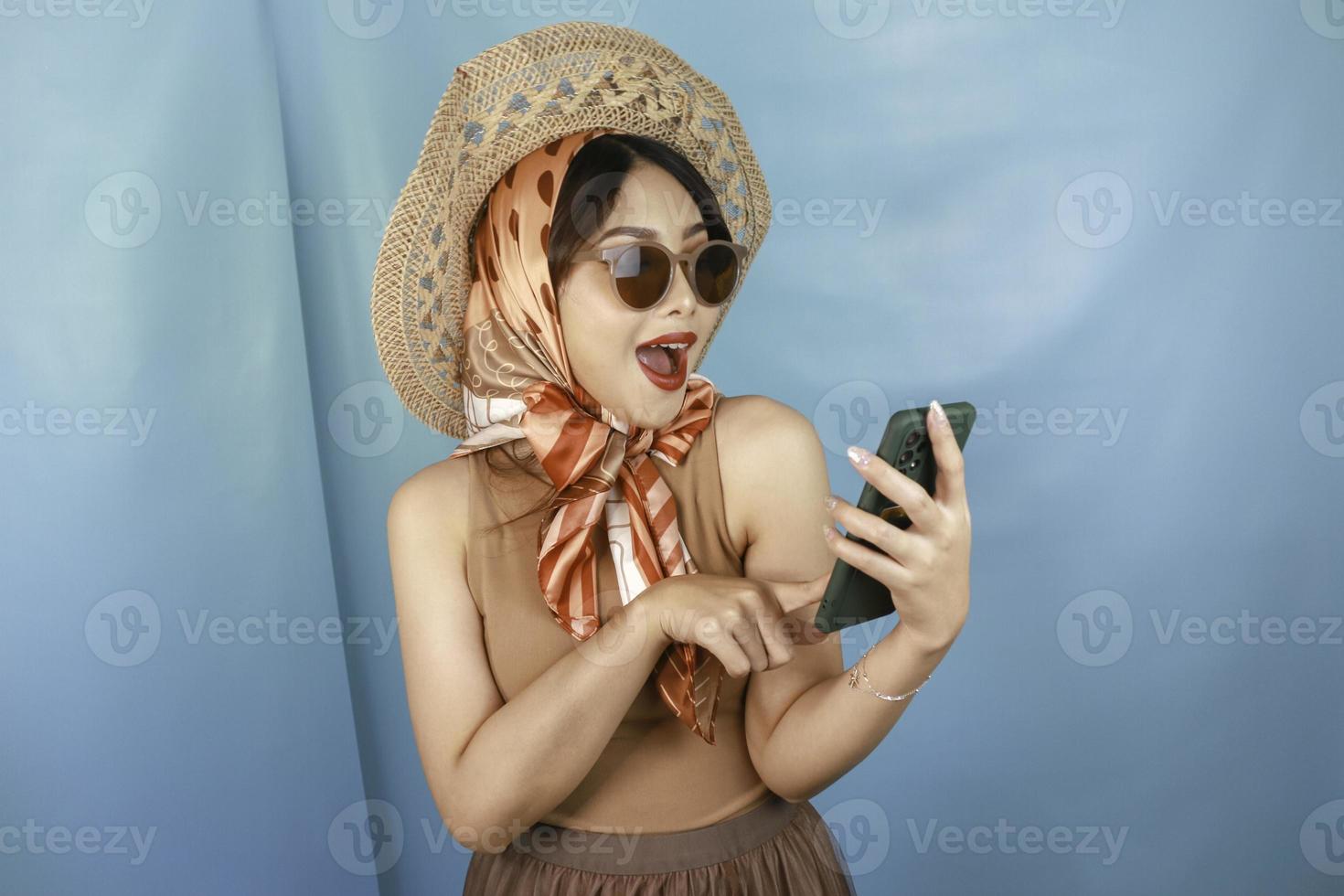 Young Asian happy woman pointing and smiling to her smartphone isolated by a blue background photo