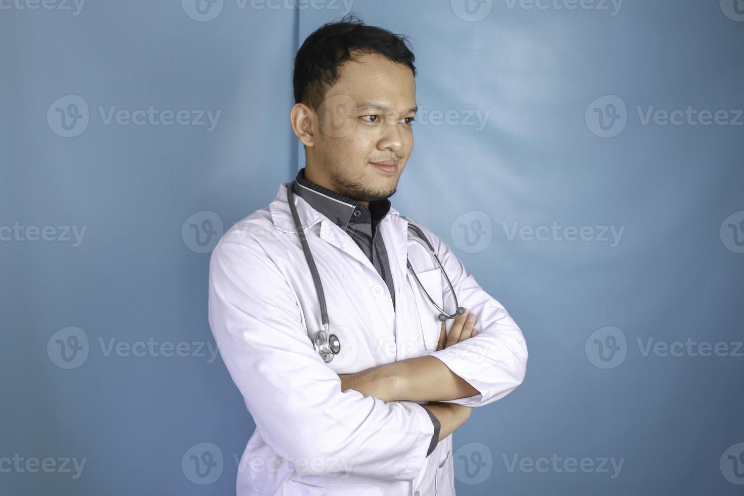 retrato de un atractivo médico varón confiado, brazos sonrientes amistosos cruzados usar bata blanca de laboratorio estetoscopio aislado fondo de color azul foto