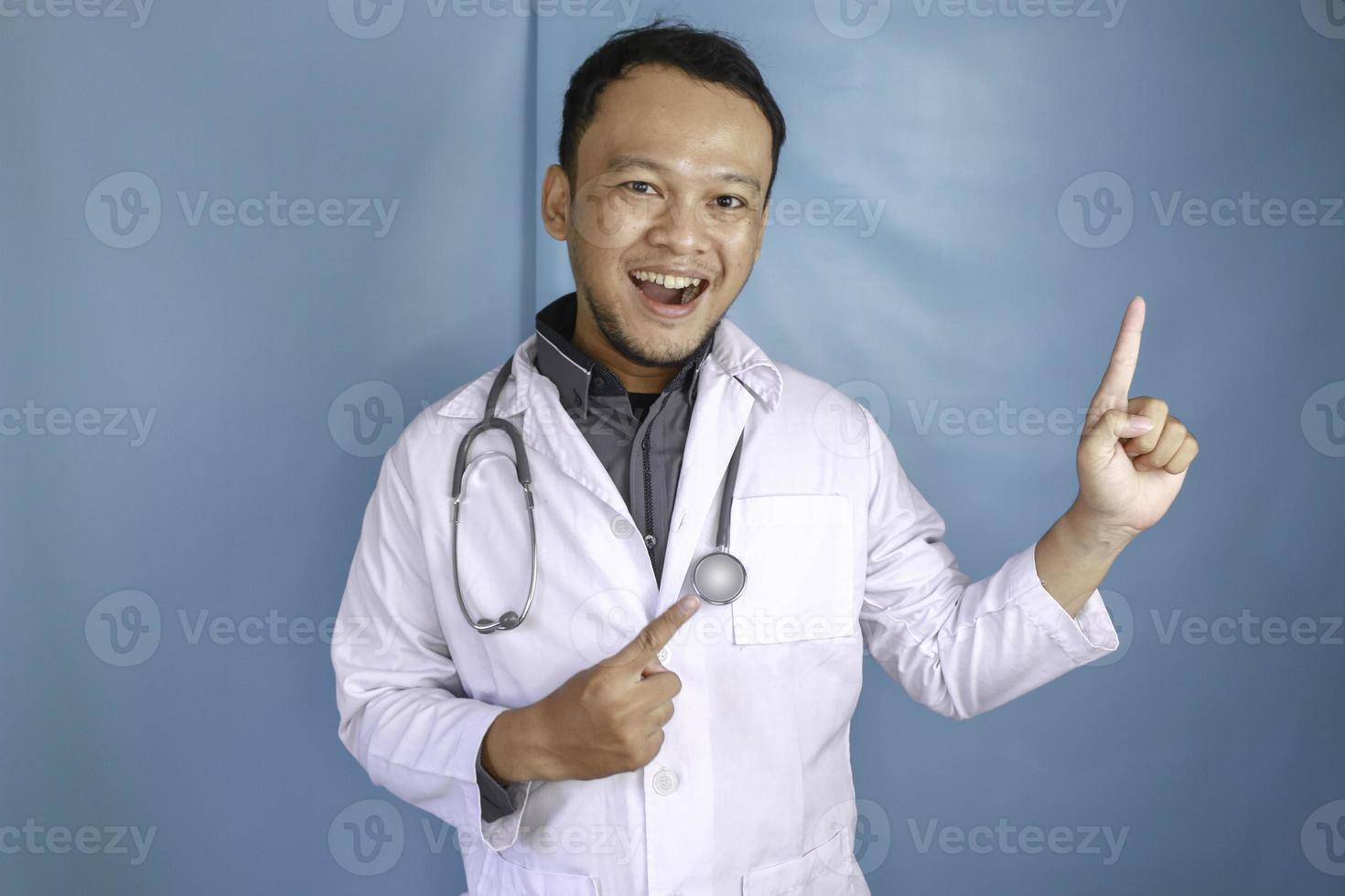 Happy young Asian man doctor, a medical professional is smiling and pointing upward at a copy space isolated over blue background photo