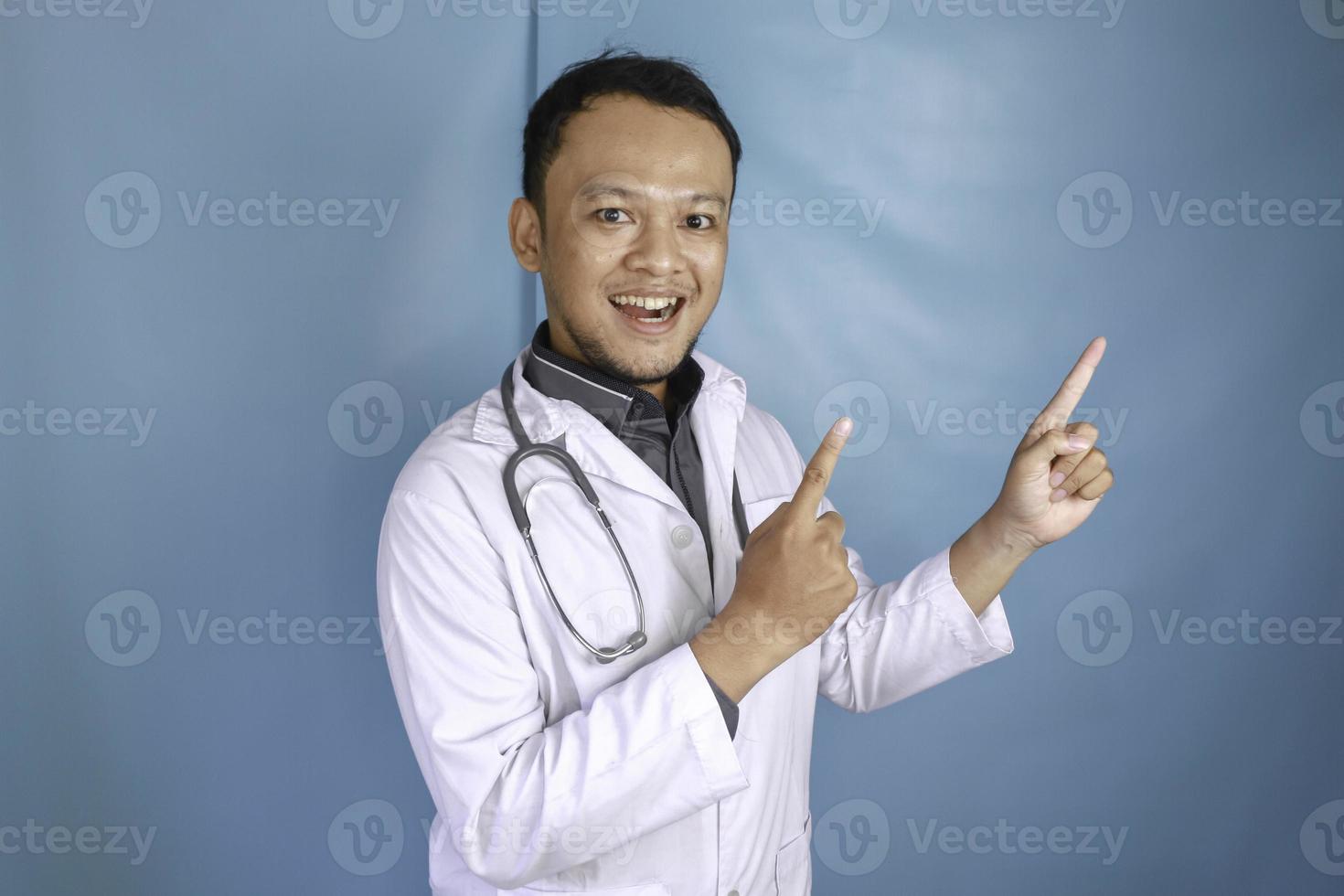 feliz joven médico asiático, un profesional médico sonríe y señala hacia arriba en un espacio de copia aislado sobre fondo azul foto