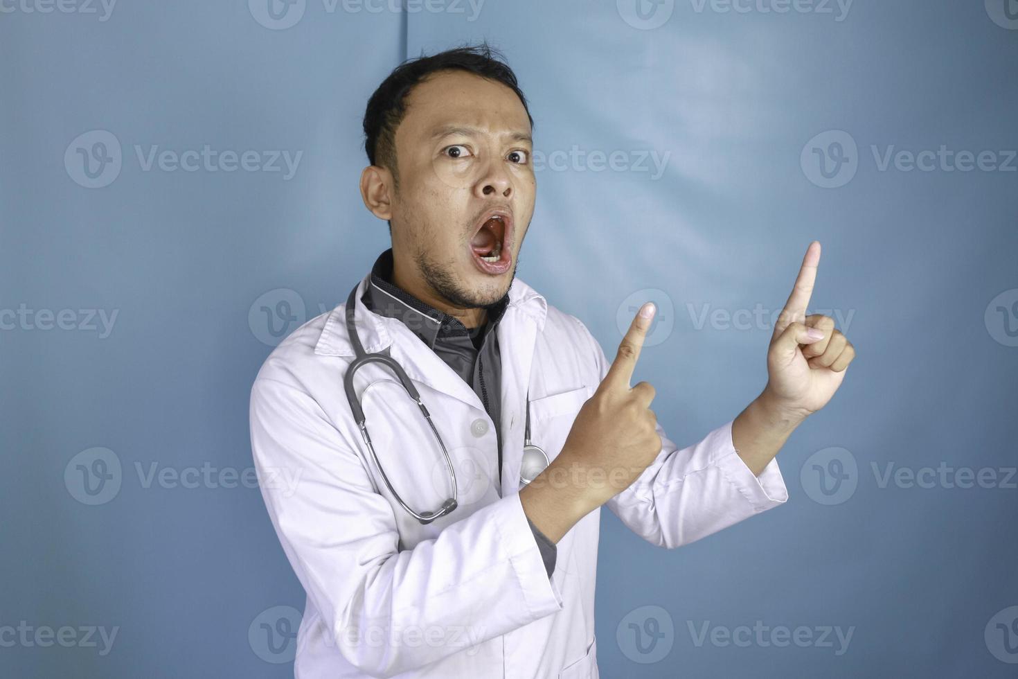 Young Asian man doctor, a medical professional is shock and pointing upward at a copy space isolated over blue background photo