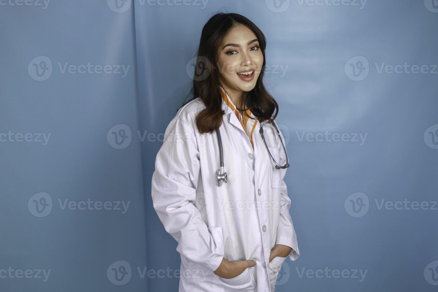 Confident relaxed female Asian doctor standing with hands in the pockets of her lab coat smiling quietly at the camera over blue background with copy space photo