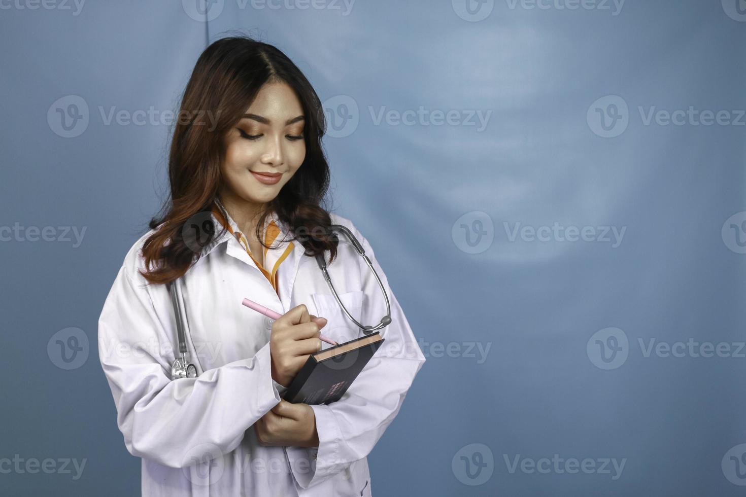 Portrait of a young Asian woman doctor, a medical professional is smiling and holding notes isolated over blue background photo