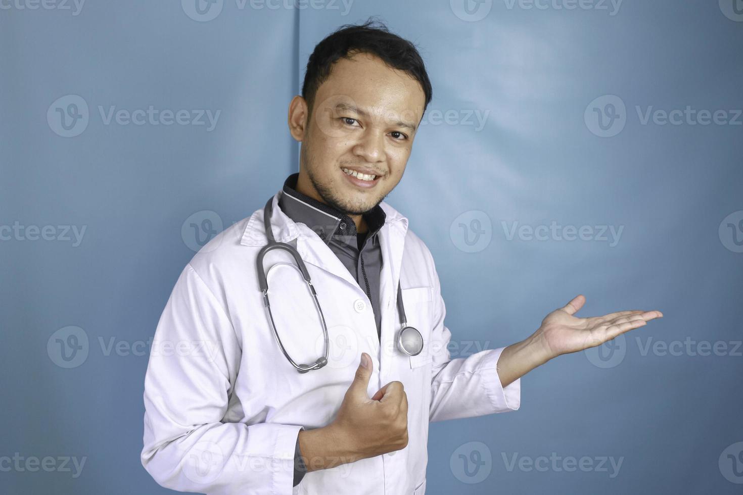 feliz joven médico asiático, profesional médico sonríe y señala un espacio de copia aislado sobre fondo azul foto