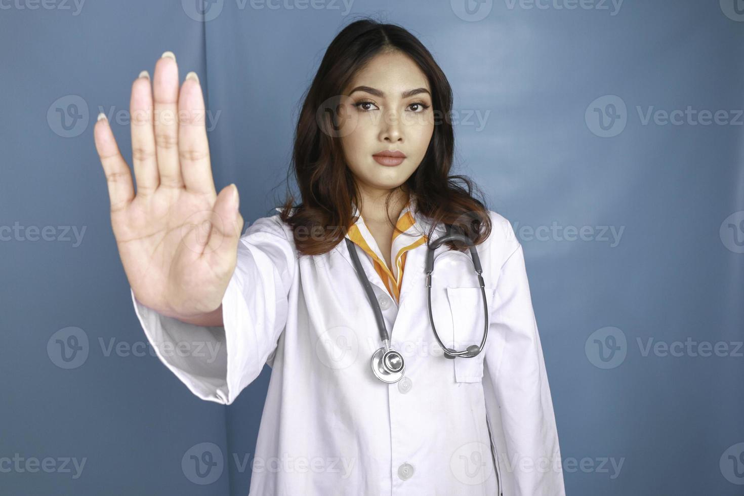 Una doctora asiática seria con estetoscopio y bata blanca, que muestra la señal de stop. foto