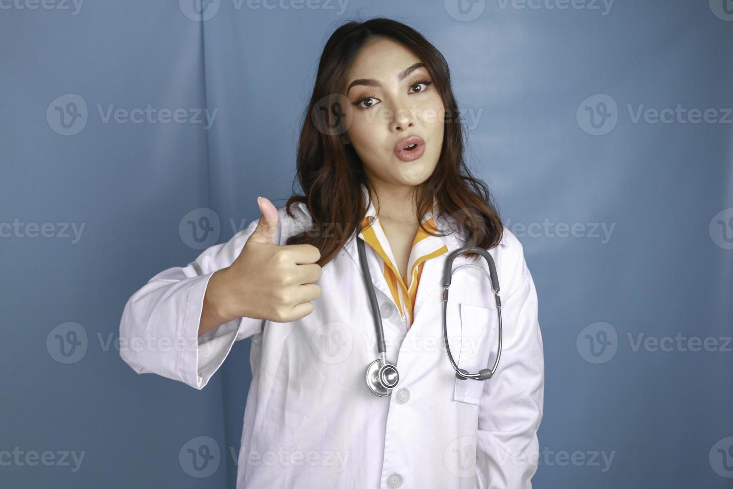 Portrait of a young Asian woman doctor, a medical professional is smiling and showing thumbs up or OK sign isolated over blue background photo