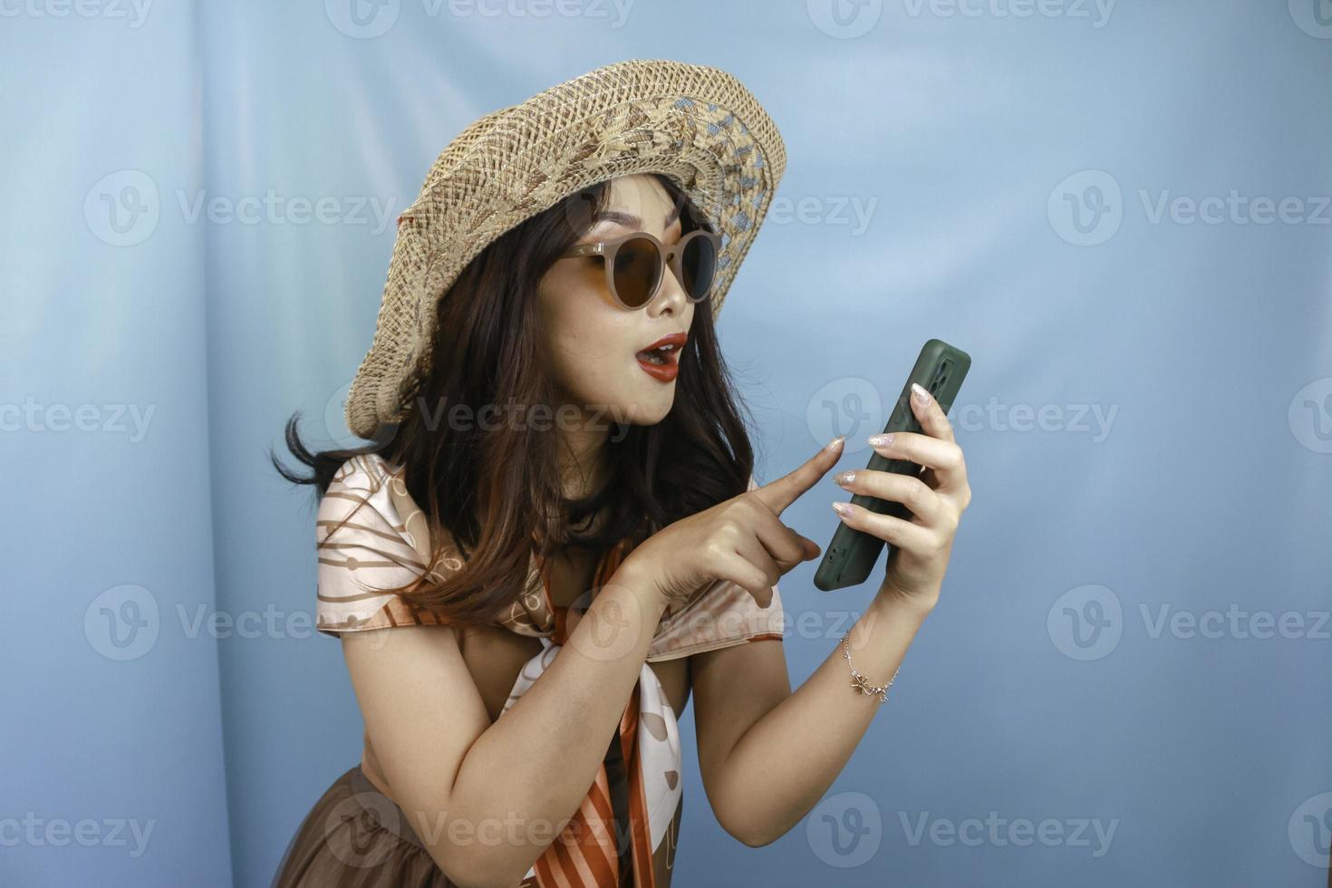 Young Asian happy woman pointing and smiling to her smartphone isolated by a blue background photo