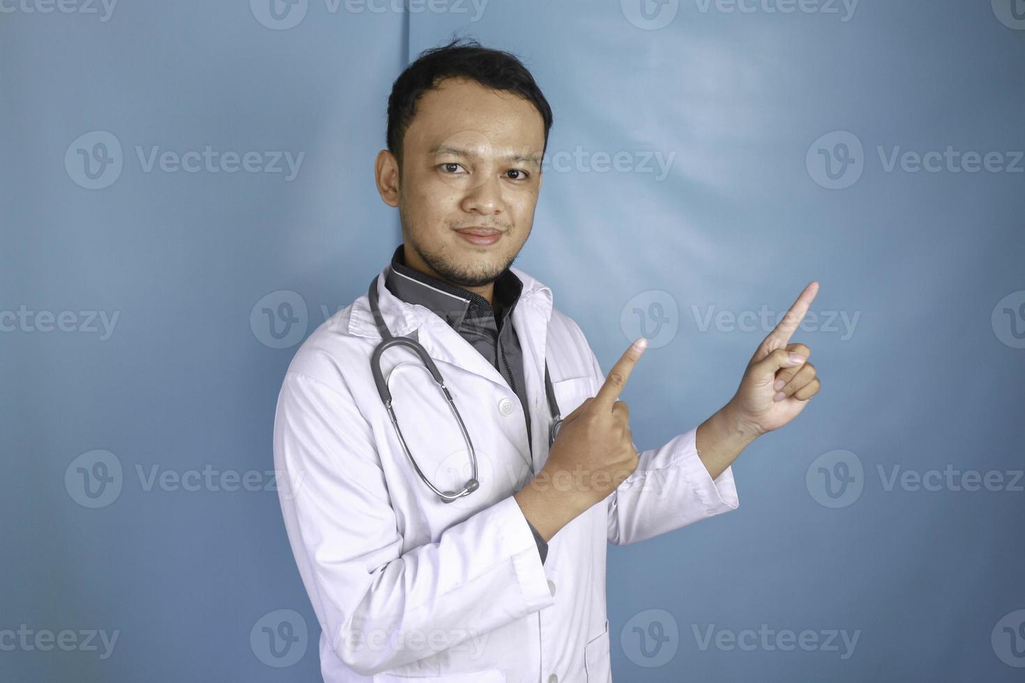 Happy young Asian man doctor, a medical professional is smiling and pointing upward at a copy space isolated over blue background photo