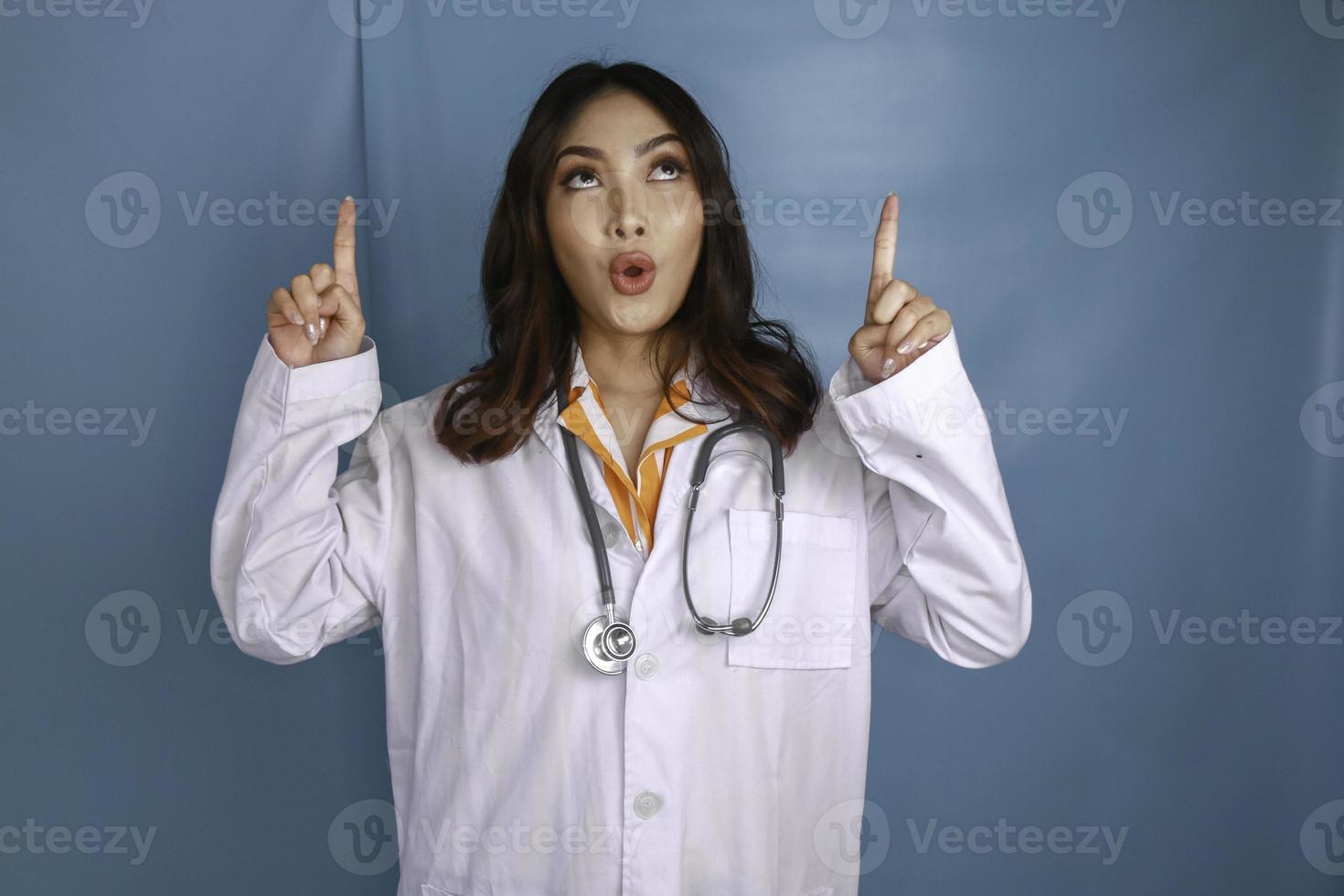 retrato de una joven doctora asiática, una profesional médica está conmocionada y apunta hacia arriba a un espacio de copia aislado sobre fondo azul foto