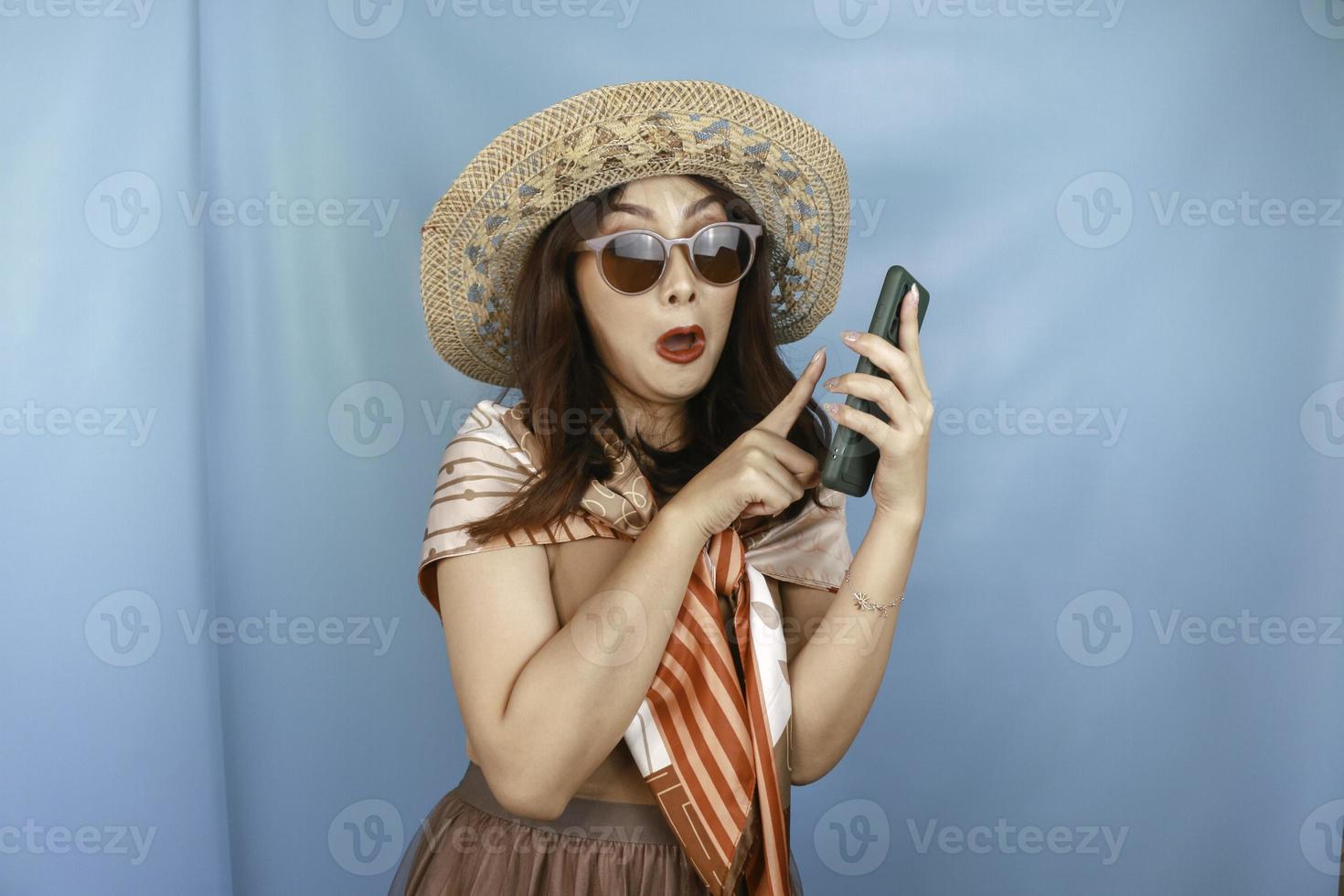 Young Asian woman pointing and shocking to her smartphone isolated by a blue background photo