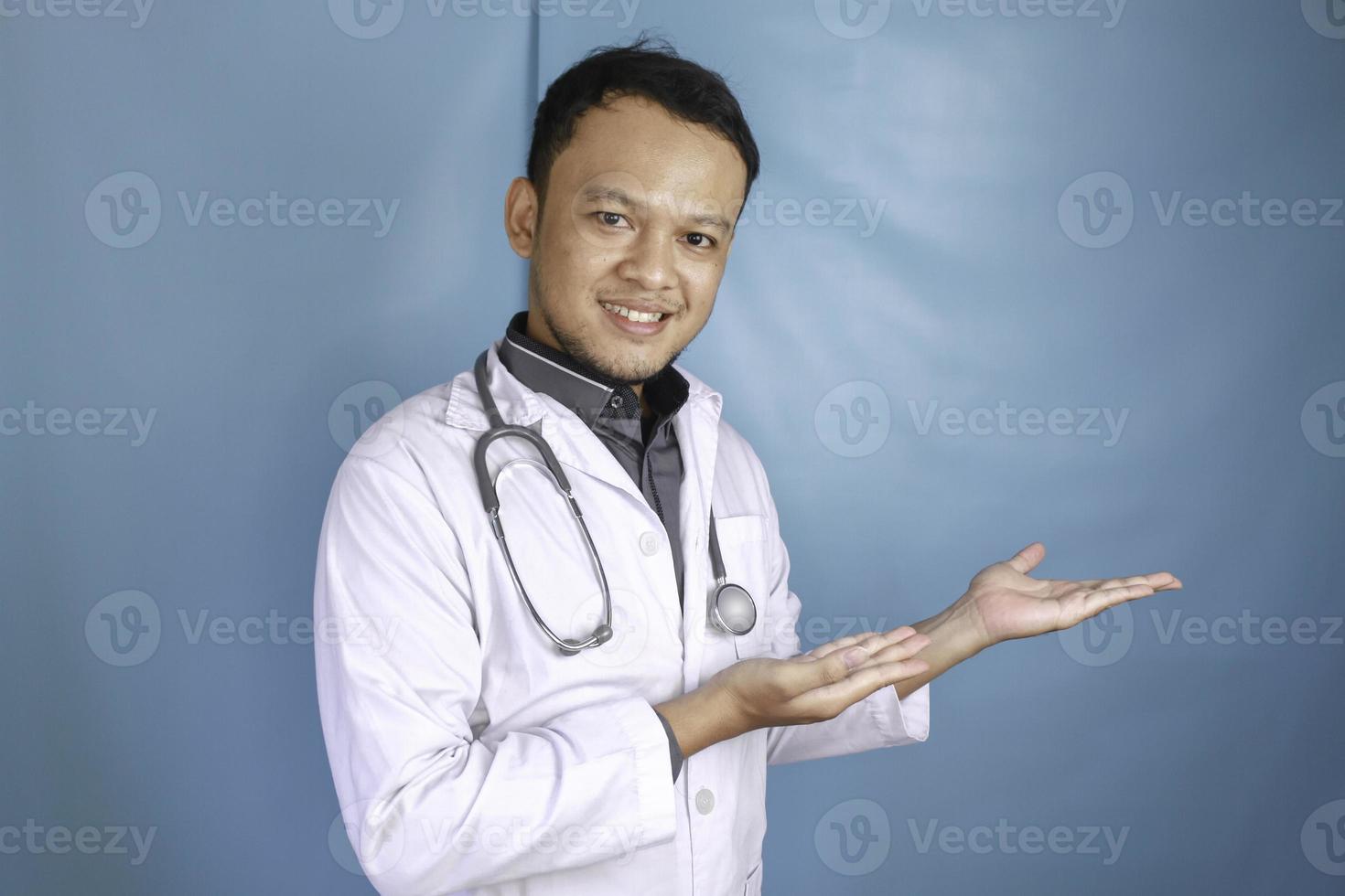 feliz joven médico asiático, profesional médico sonríe y señala un espacio de copia aislado sobre fondo azul foto