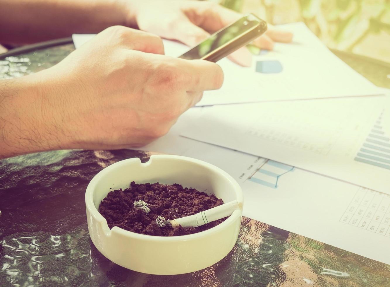 A businessman is seriously working with mobile phone looking at graph paper with cigarette butt in ashtray, selective focus of business and financial working concept. photo