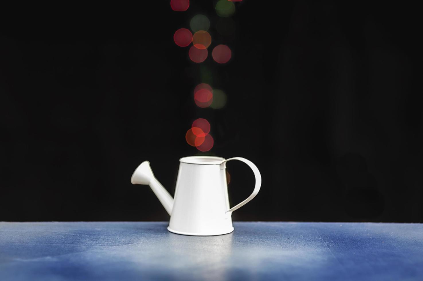 Abstract photo of small decorative white watering can putting on blue table with colorful steam bokeh going up over black background