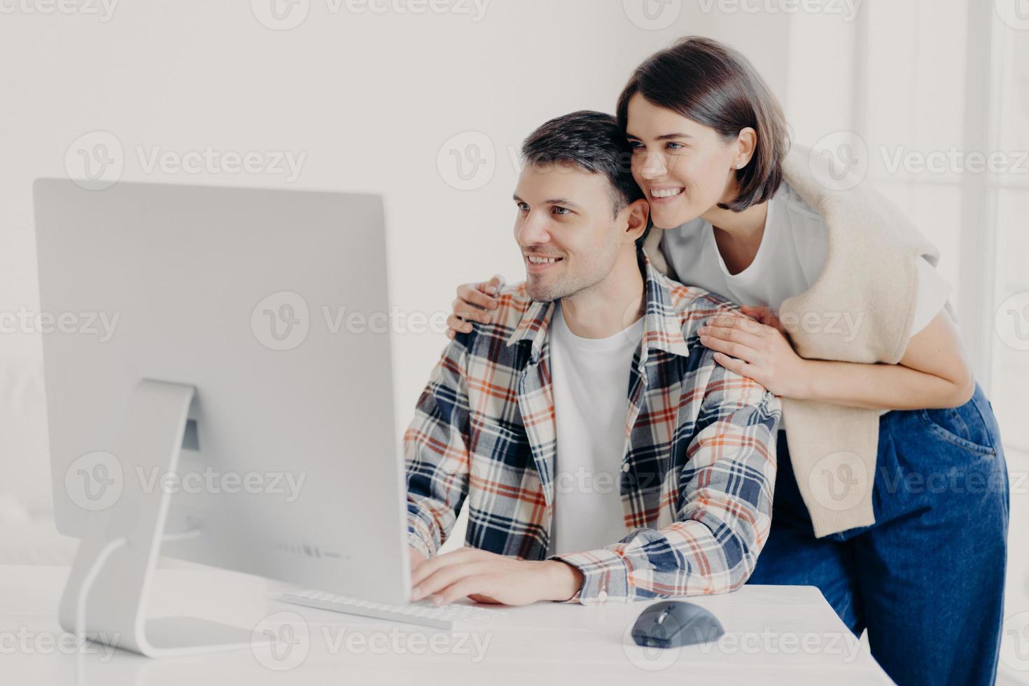 Young couple cooperate for developing website, work on common internet project, affectionate wife hugs husband with support, focused into monitor, keyboard information, connected to internet photo
