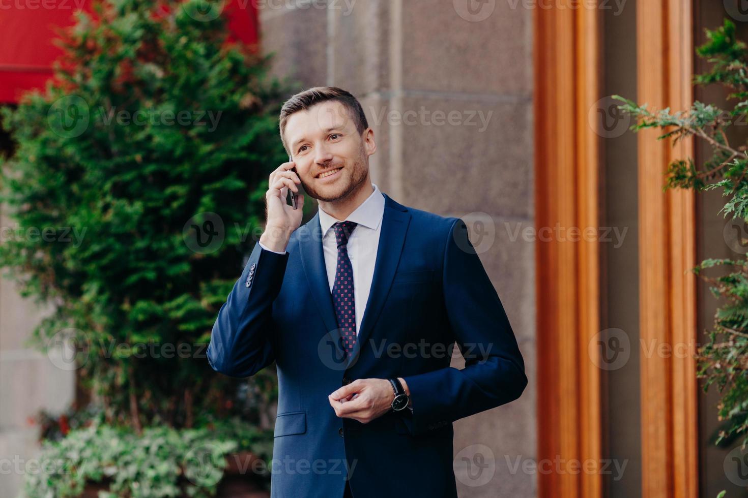 Successful businessman has work break, uses mobile phone for having talk, being always on line, dressed in formal suit, has pleasant smile on face, stands outdoor near restaurant. Technology concept photo