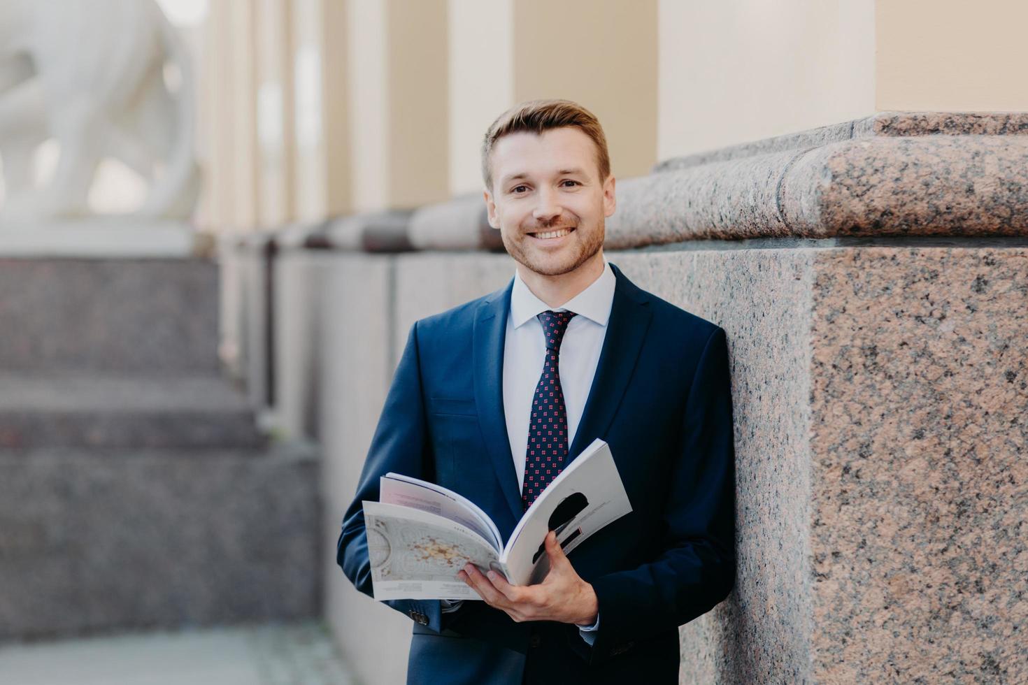 Prosperous successful businessman in formal outfit holds journal, has happy expression, smiles gladfully, enjoys dinner break after hard working day, being in good mood after business meeting photo