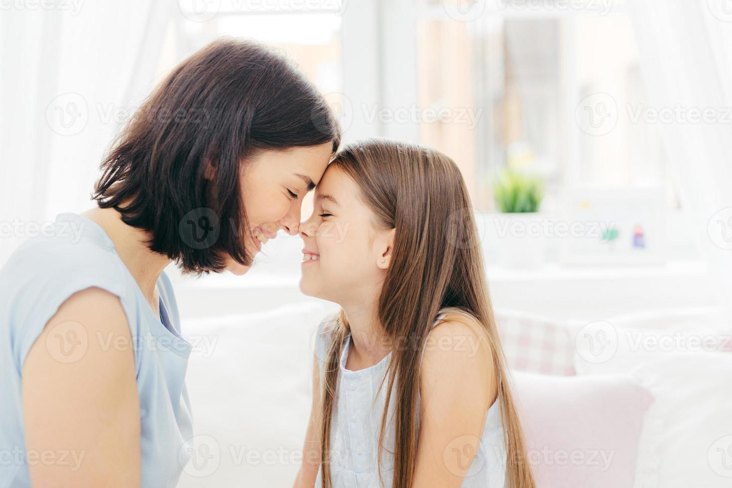 Close up shot of affectionate young mother and her little daughter touches noses and enjoy togetherness, pose against white background and bedroom interior, smile gladfully. Motherhood concept photo