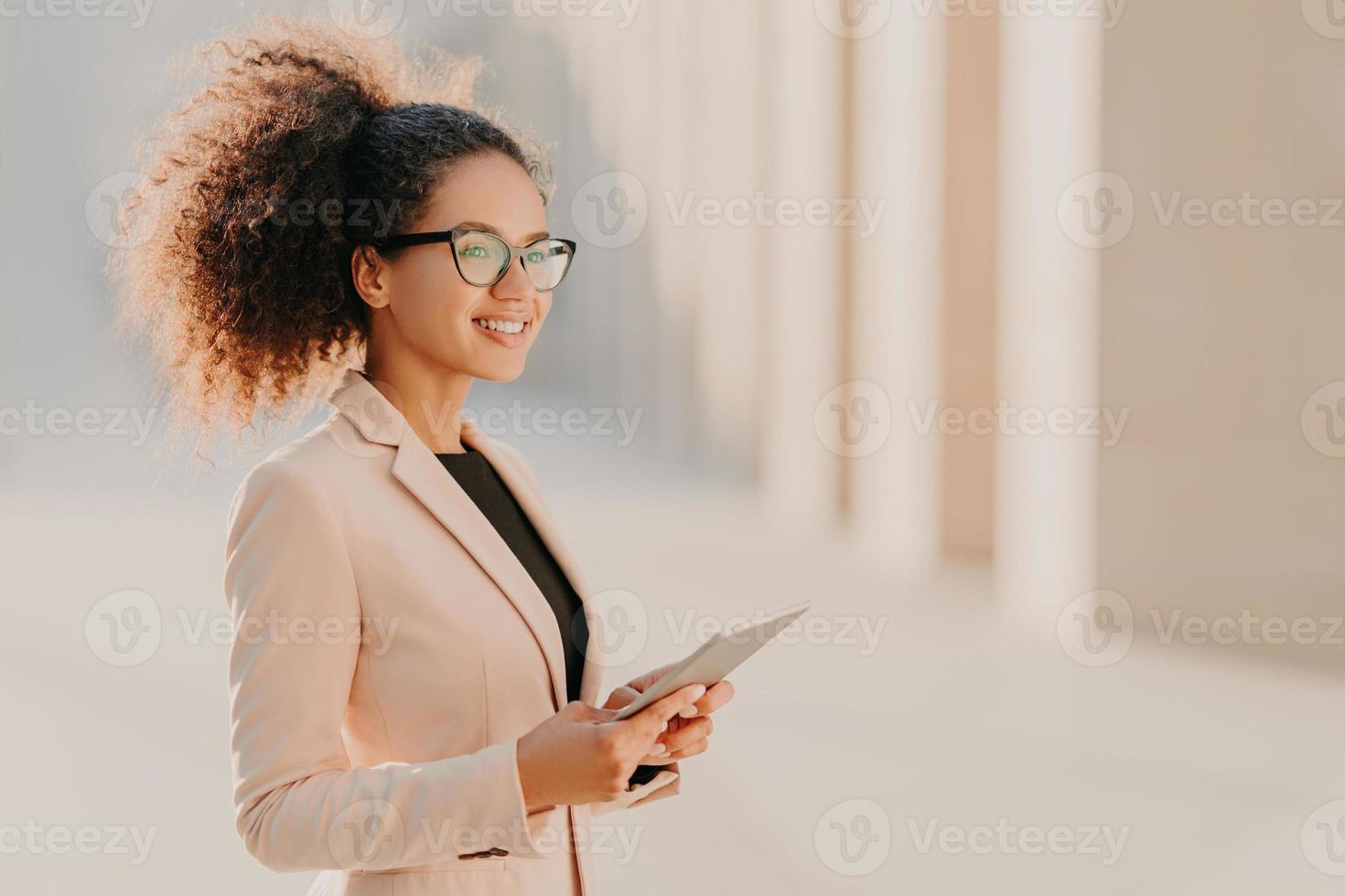 Profile shot of glad dark skinned woman with Afro hair, dressed elegantly holds tablet computer in hands strolls at street wears optical glasses connected to high speed internet. Lifestyle, technology photo