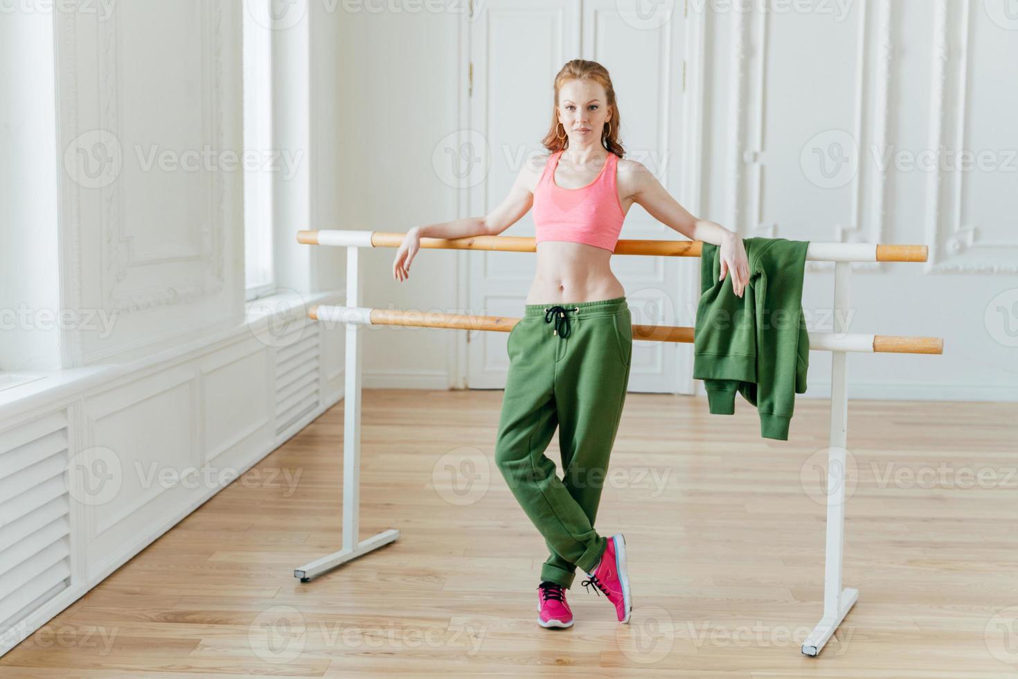 Slim beautiful female ballet dancer leans at ballet barre, wears pink top, sport trousers and sneakers, shows fit figure, stretches before dancing, poses in studio or classroom, has rehearsal photo