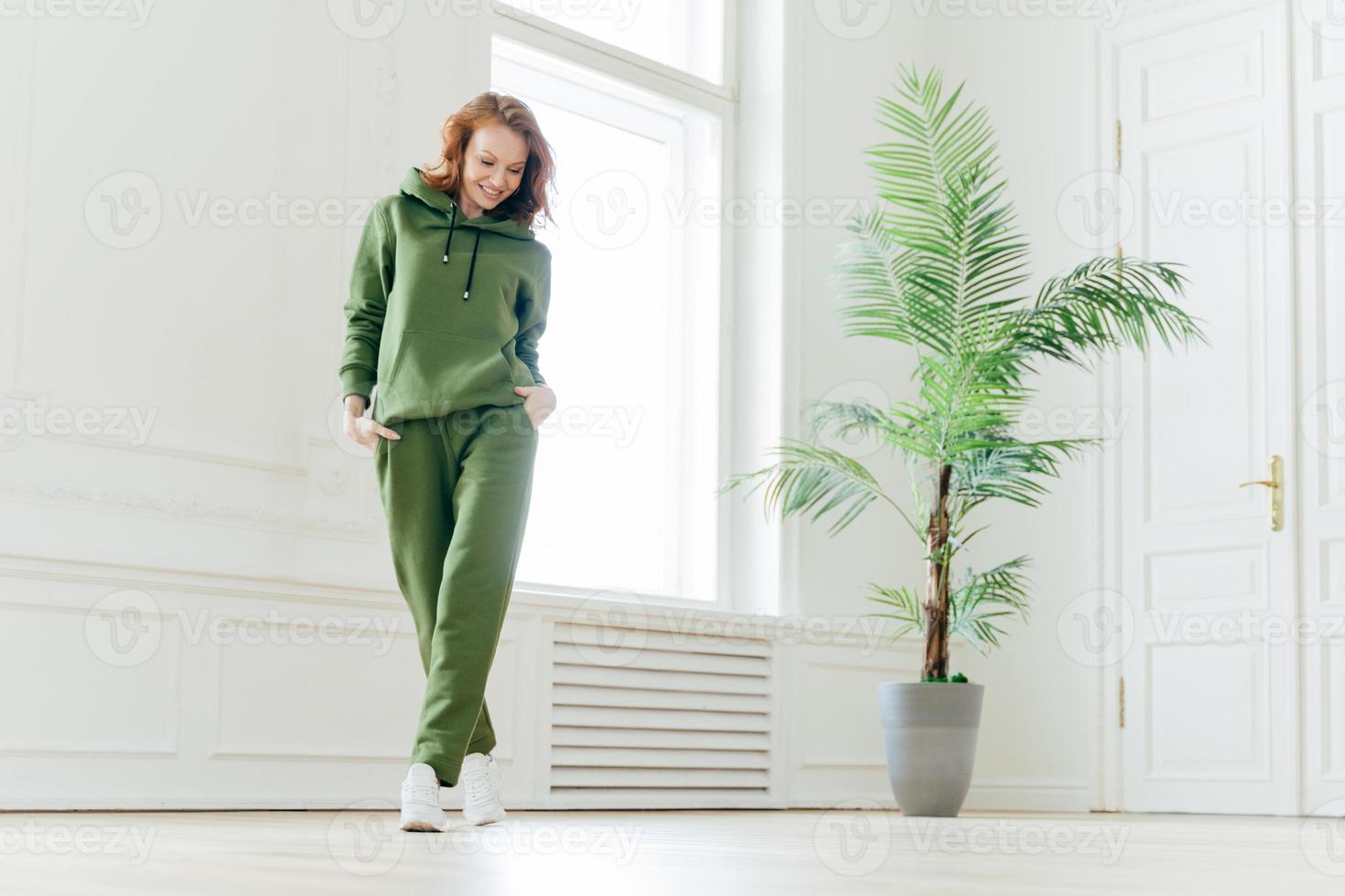 Full length shot of active pleased young woman with slim body, keeps hands in pockets of tracksuit, focused down, ready for doing cardio exercises, has determined look, pilates exercising indoor photo