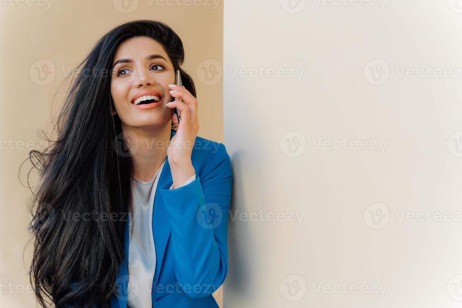 Young positive businesswoman smiles happily, communicates on mobile phone, dressed formally, focused somewhere, has minimal makeup. Cheerful female entrepreneur solves working issues in good mood photo