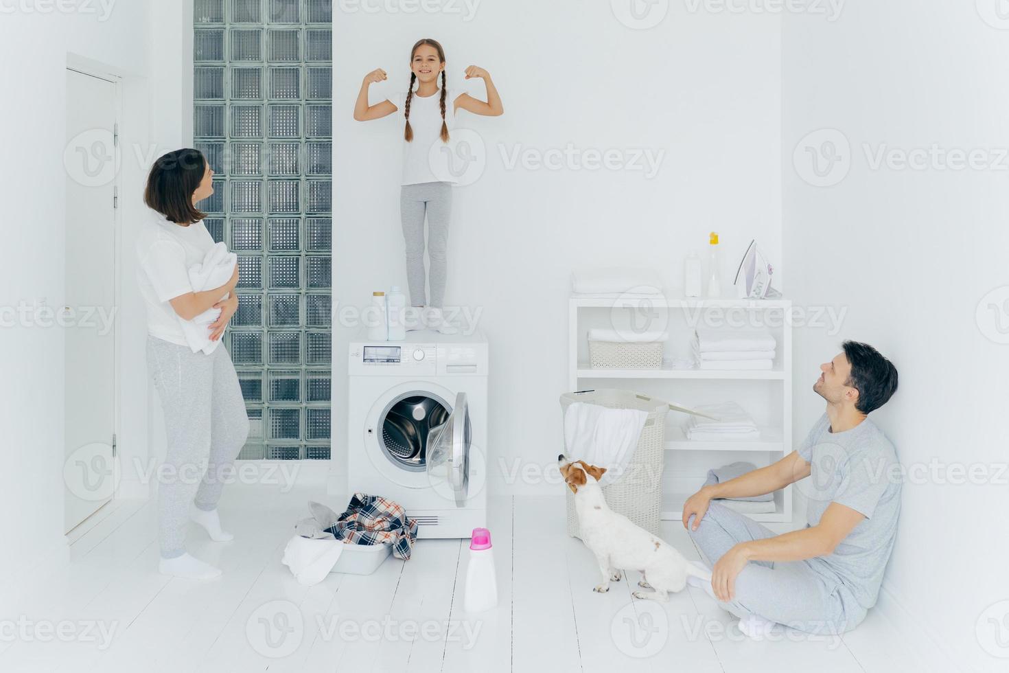 Photo of happy schoolgirl poses on top of washing machine, shows muscles, raises arms, ready to help parents with washing or laundry. Woman and man pose in washing room with dog and small kid