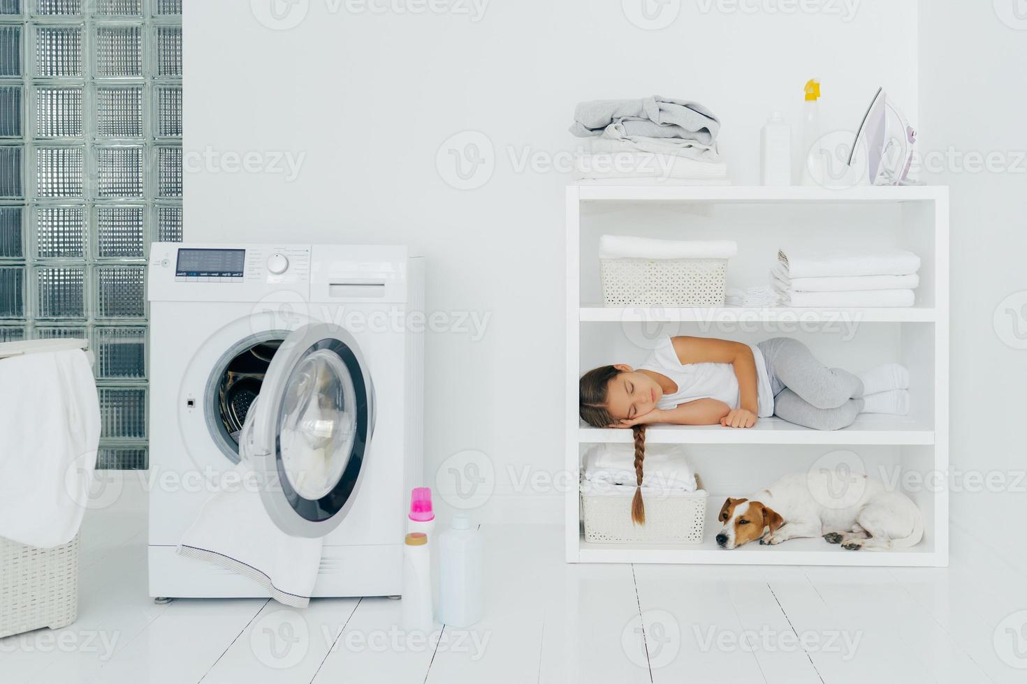 Tired female child rests on console shelf together with pet, does washing at home, sleeps in laundry room, opened washing machine with dirty towel inside. Childhood, cleanliness, family chores concept photo