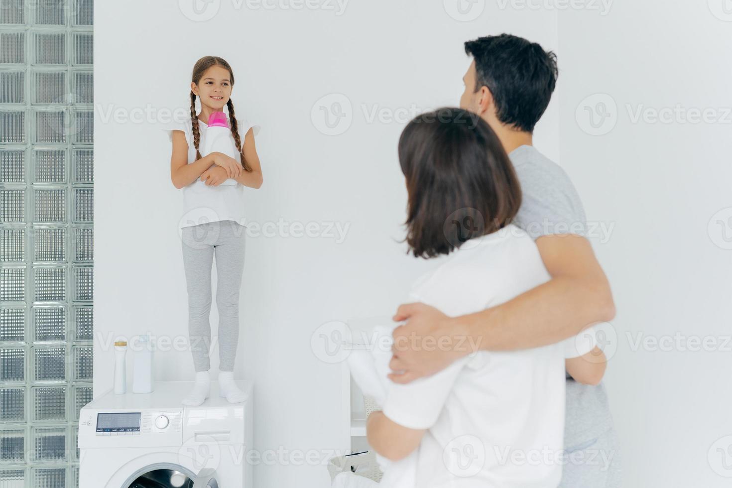 Back view of husband and wife embrace and talk to small girl standing on washer with bottle of detergent, do washing together during day off, have much work about house. Laundry day, home routine photo