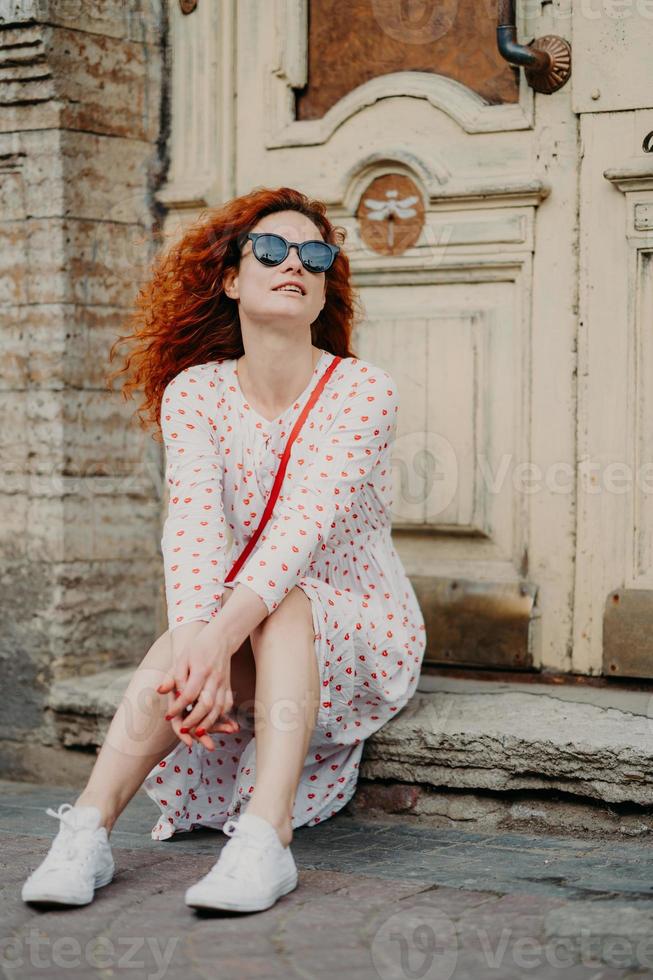 Pleased red haired female with curly hair, wears sunglasses and dress, poses near door of ancient building, holds hands together, enjoys rest in open air, likes viewing old historic famous places photo