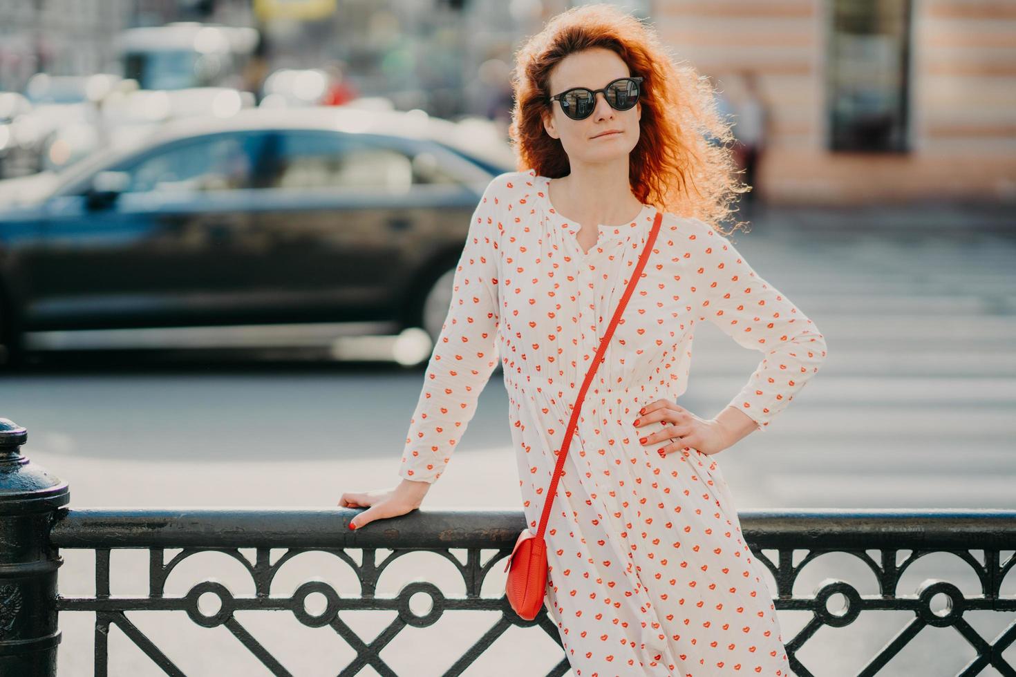 Outdoor shot of satisfied redhead woman keeps one hand on waist, other on street hence, poses over blurred urban setting, wears sunglasses and dress, has red manicure. Copy space aside for text photo