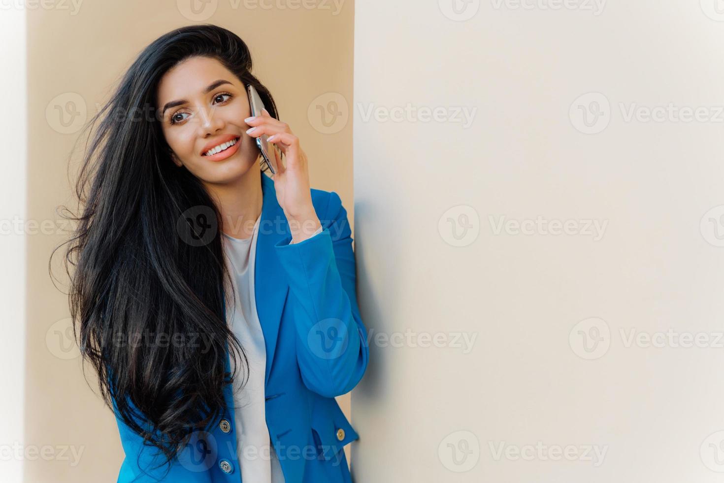 Happy female model with dark long hair calls via cell phone and talks in roaming wears blue jacket looks aside has thoughtful expression. Office worker speaks on cell phone. Business and communication photo