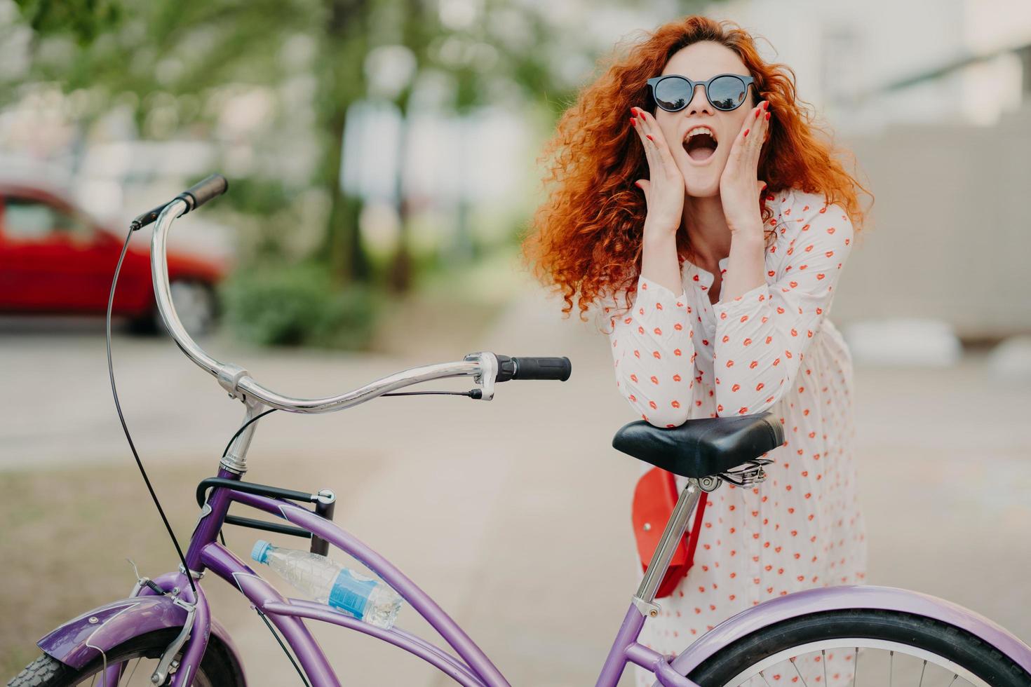Horizontal shot of happy woman rides bicycle in park, has break, poses at camera, keeps mouth opened, touches cheeks, wears trendy sunglasses, has curly red hair, enjoys nice weekend during summer photo