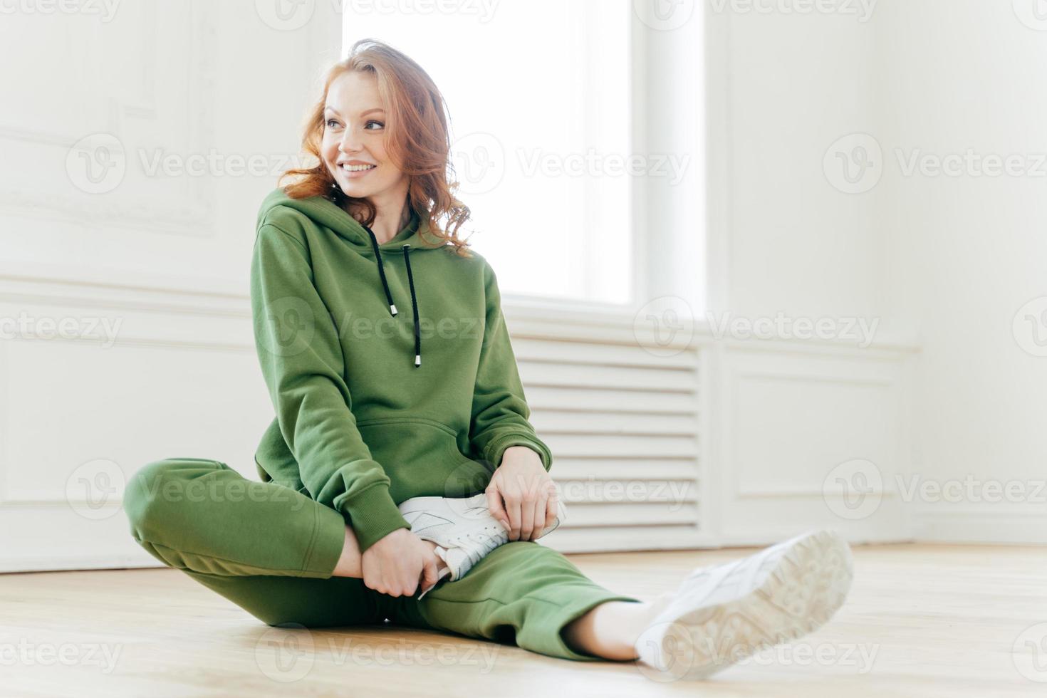 Pleased redhead woman focused aside, has happy expression, stretches legs, sits on floor, waits for instructor, wears hoody, white sneakers, has yoga exercising. People and motivation concept photo