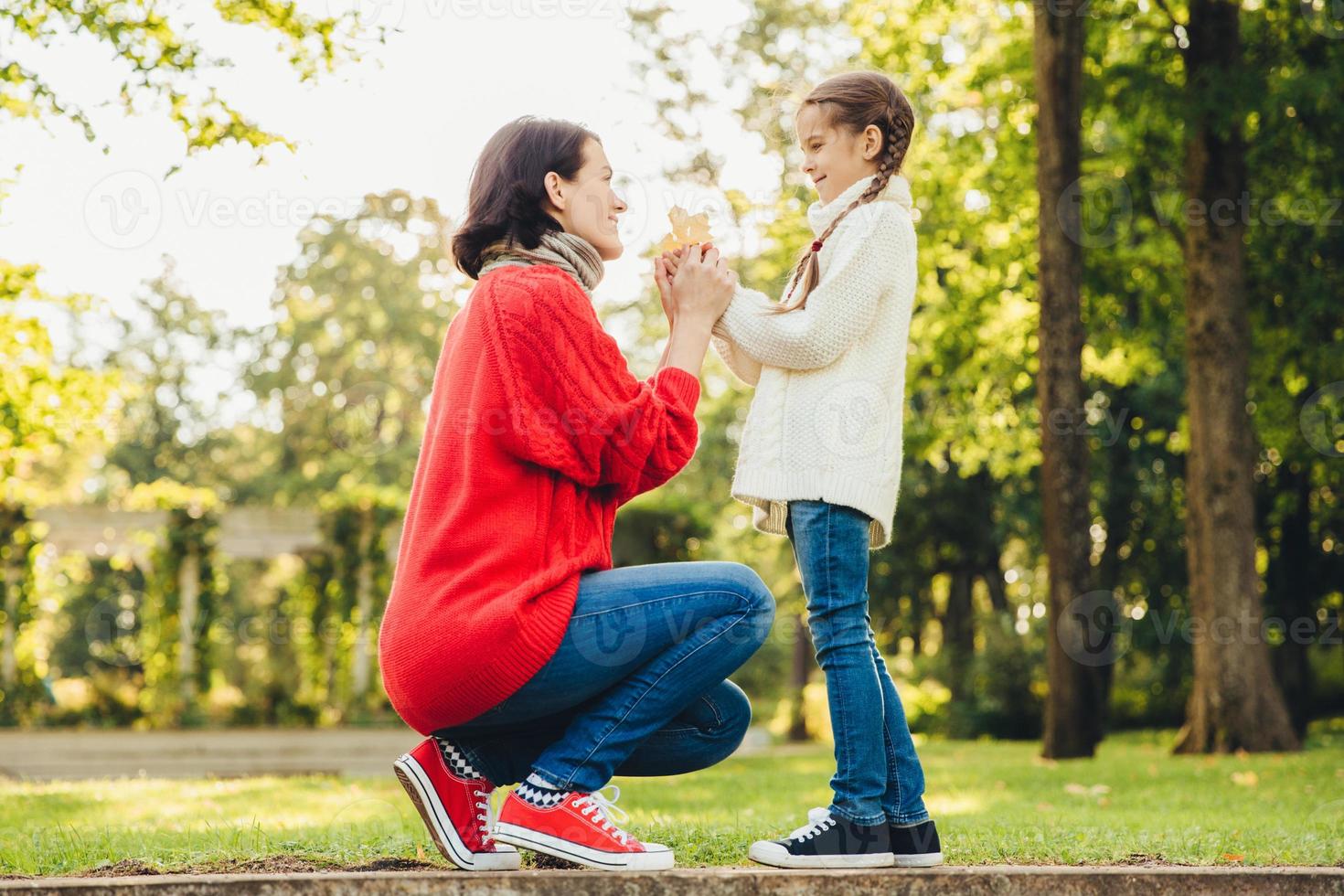 Young mother in warm knitted red sweater plays with her small daughter in park, gives her leaf, enjoy sunny autumn weather. Affectionate mom and little child spend time together outdoor photo
