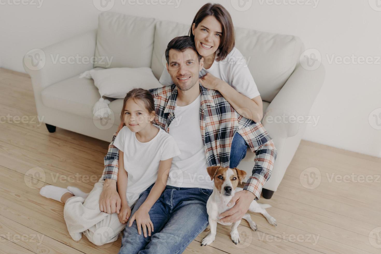 Top view of happy family members pose in spacious room near comfortable sofa, favourite dog poses near, enjoy spare time sit on floor, smile positively. Father, mother and child spend time at home photo