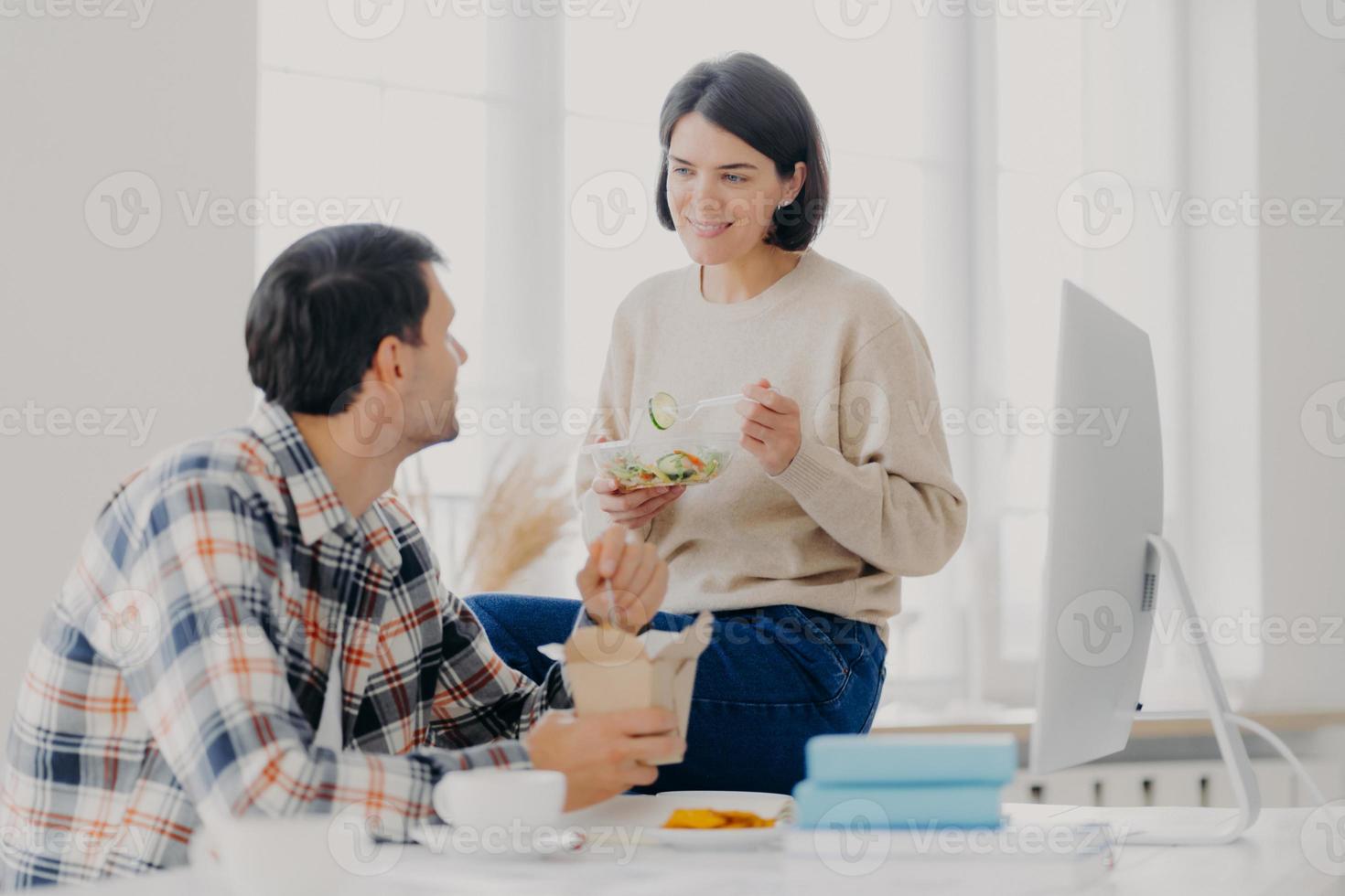 Photo of lovely woman and man discuss working issues together while have lunch, eat fast food and fresh vegetable salad, pose at desktop with computer monitor and scientific literature. Teamwork