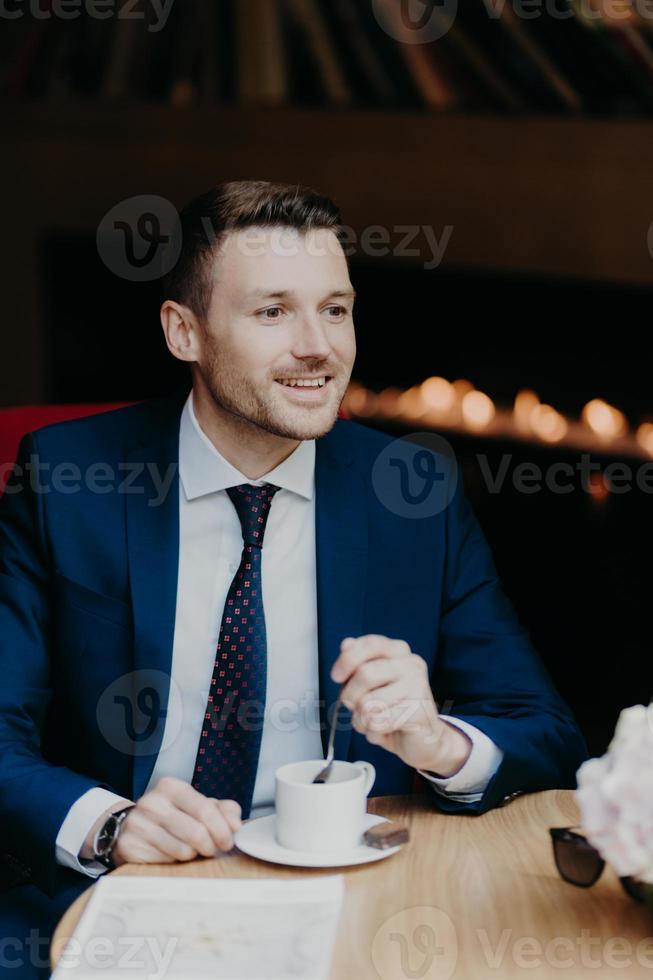 Vertical shot of handsome male in formal clothing, has cheerful expression, has dinner break at cafeteria, drinks aromatic coffee, looks happily somewhere, thinks about something. Business and rest photo