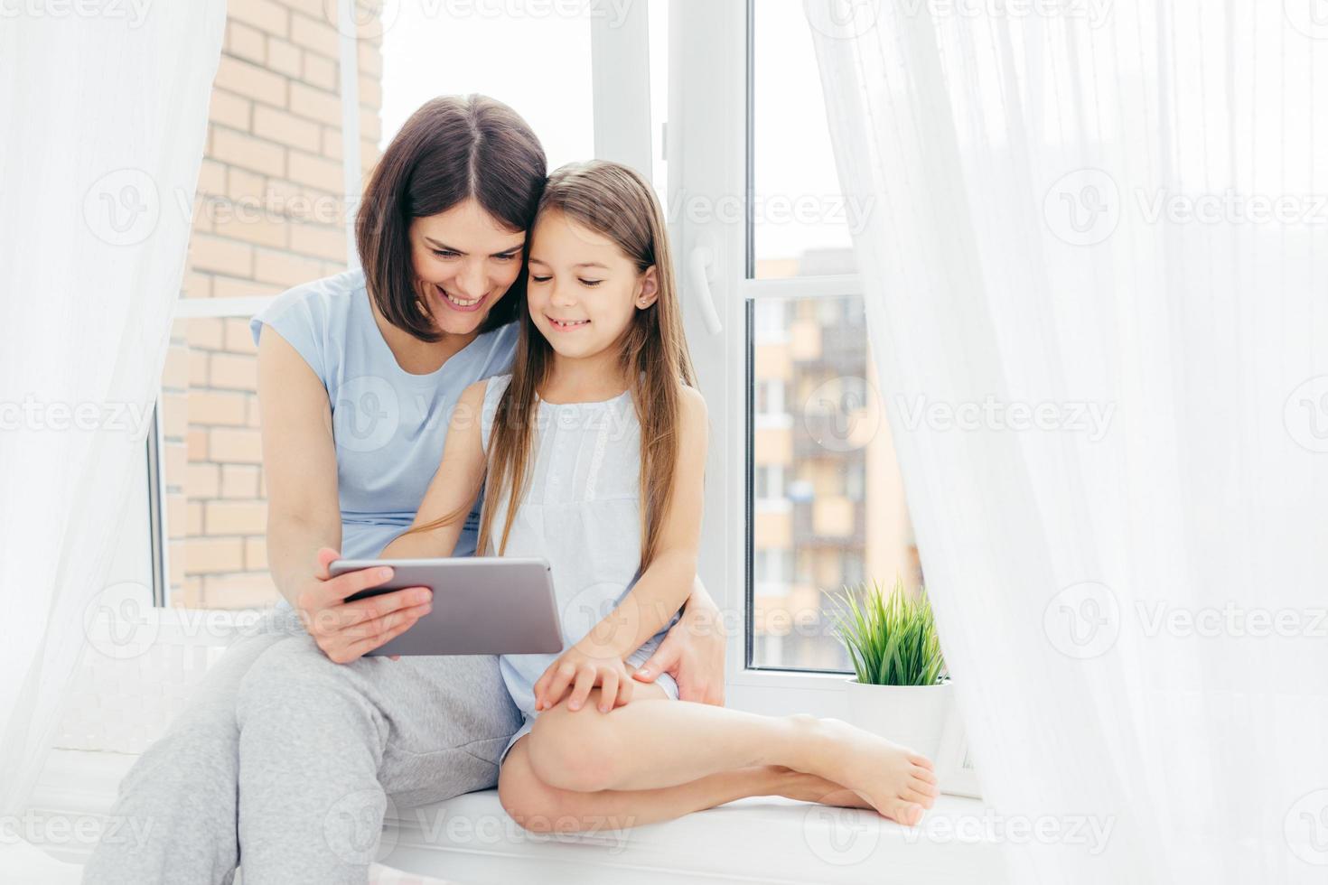 People, technology, family, children concept. Positive young other and her small daughter sit on window sill, hold digital tablet, watch interesting cartoon, pose near window with white curtains photo