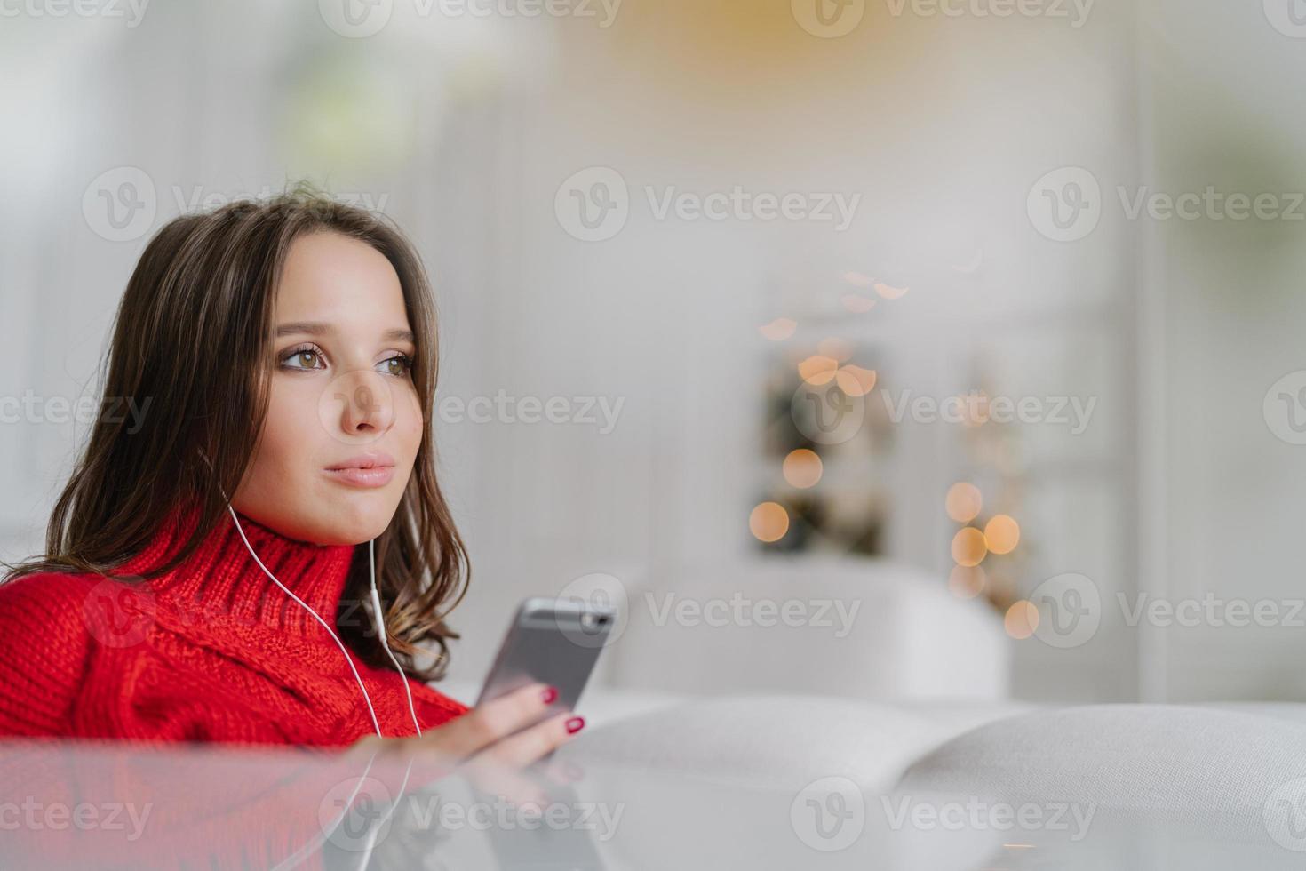 Sideways shot of thoughtful Caucasian woman dressed in knitted sweater, holds modern cell phone in hands, listens audio with earphones, connected to wireless internet, poses in living room alone photo