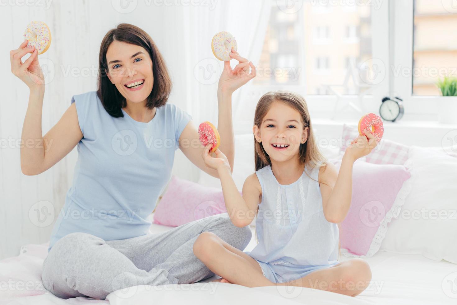 madre e hija divertidas vestidas con ropa de dormir, tienen buen humor por la mañana, toman deliciosos donuts, van a desayunar postre, posan juntos en el dormitorio. concepto de familia y unión foto