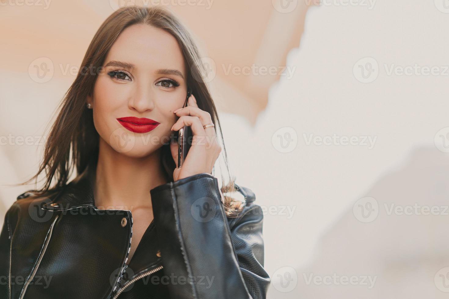 atractiva y encantadora mujer disfruta de tarifas en roaming, habla por teléfono móvil con su mejor amiga, se viste con ropa de moda, usa lápiz labial rojo, maquillaje, se para en el fondo borroso de algún edificio foto