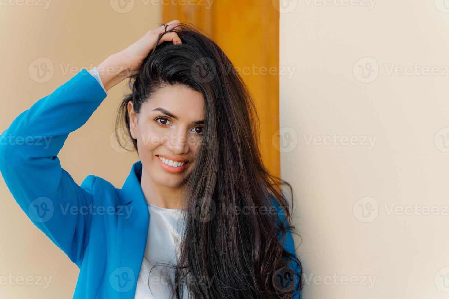 Headshot of happy brunette woman with gentle smile, makeup, wears elegant apparel, glad after successful business meeting, looks at camera, keeps hand on hair. Glad lady poses in formal clothes photo