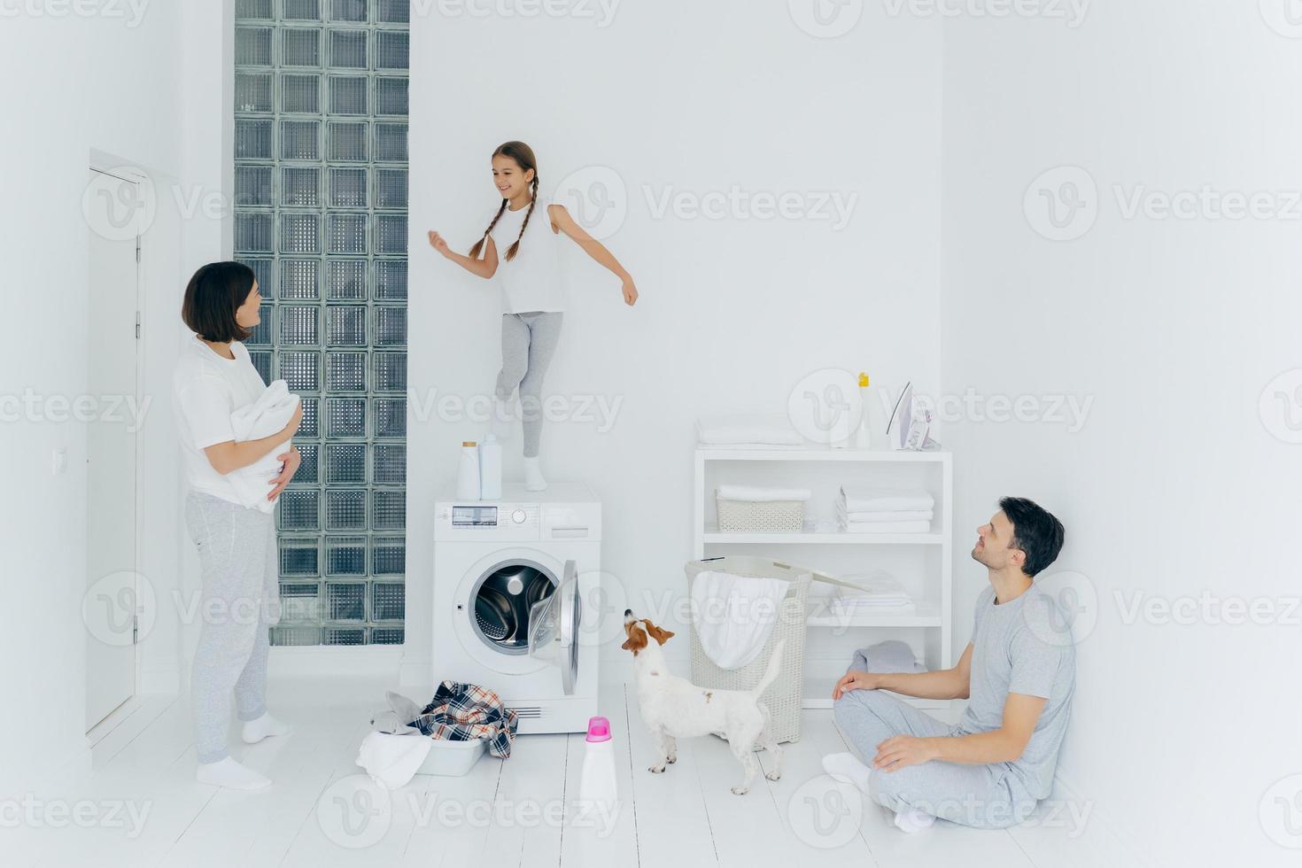 la familia feliz lava la ropa en casa, el padre se sienta en el suelo en posición de loto, la madre se para con una toalla blanca, mira al niño que baila alegremente en la lavadora, el perro pedigrí cerca. tareas del hogar foto