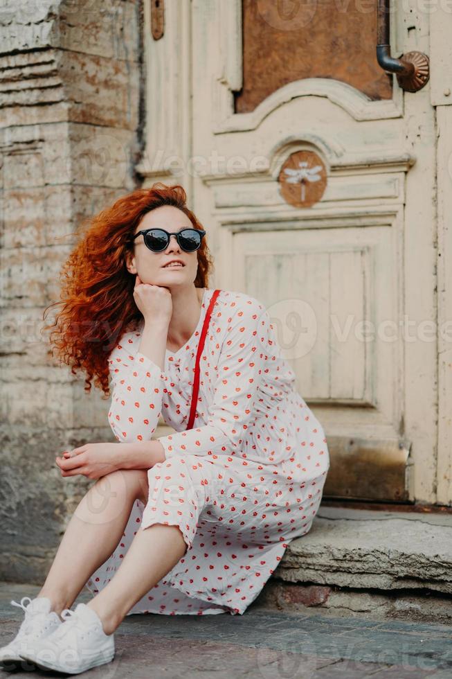 Red haired thoughtful woman has rest after strolling, poses near old building, sits on threshold, wears sunglasses, dress and white sneakers, explores ancient places in European city. Vertical shot photo