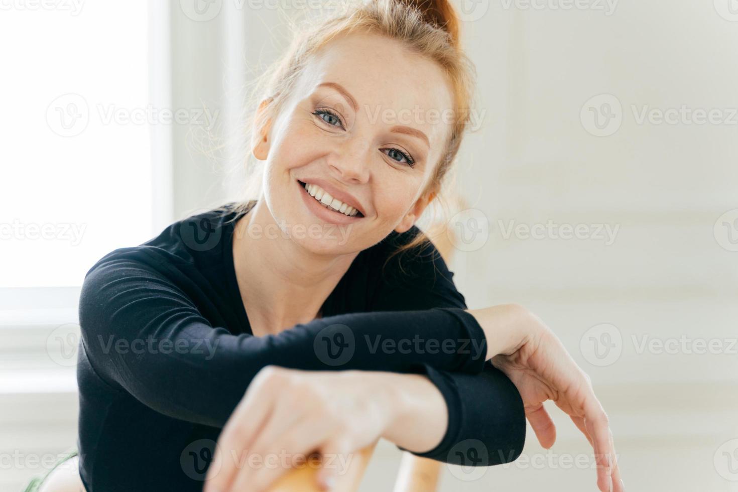 Carefree good looking ginger young female ballerina leans at ballet barre, looks gladfully at camera, practices different dancing positions, has toothy broad smile, white teeth, exercises indoor photo