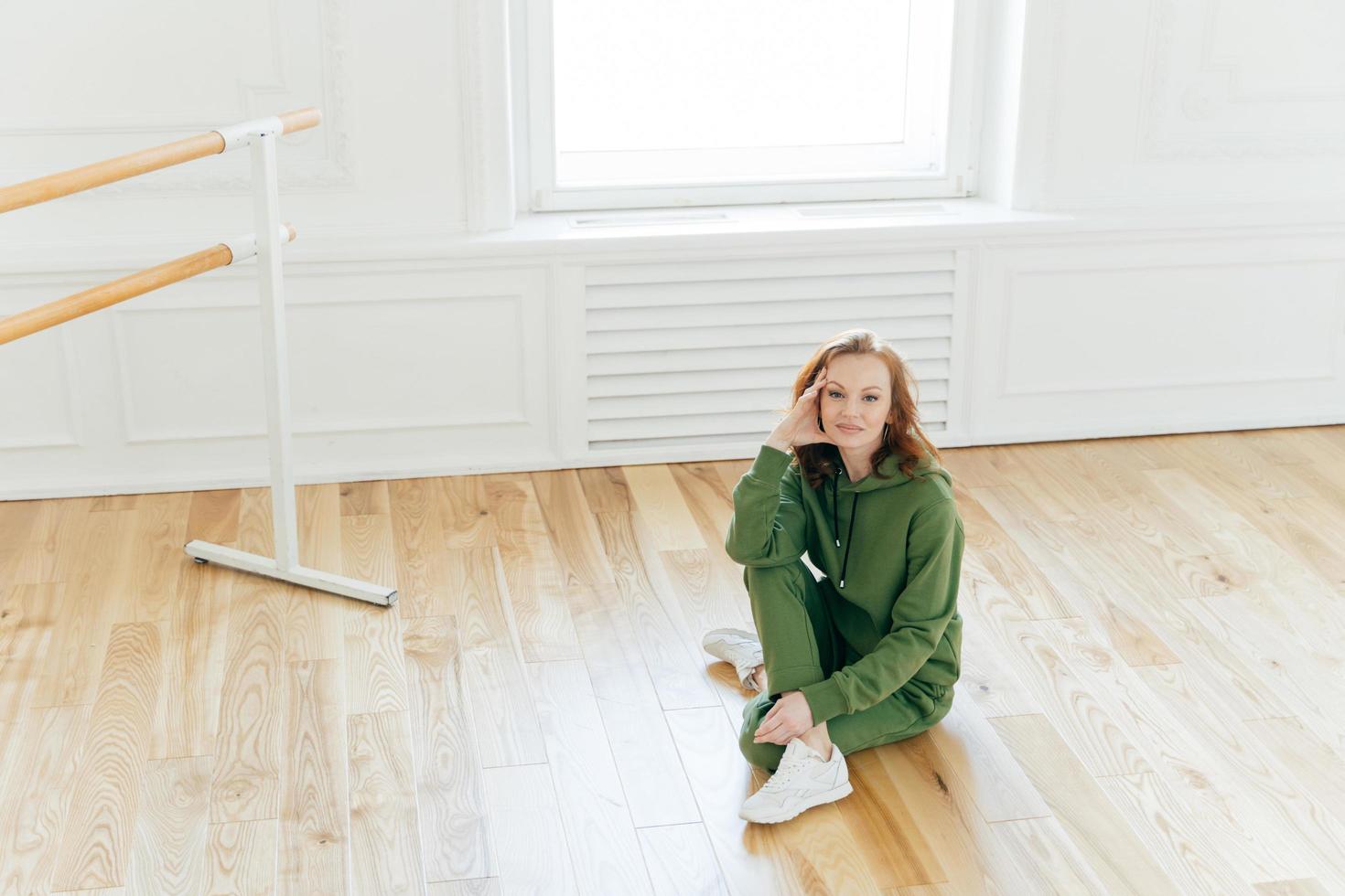 Indoor shot of satisfied good looking redhead female sits on floor, keeps legs crossed, ballet barre stands near, being professional dancer, wears sweatsuit, does stretching, poses in rehearsal room photo