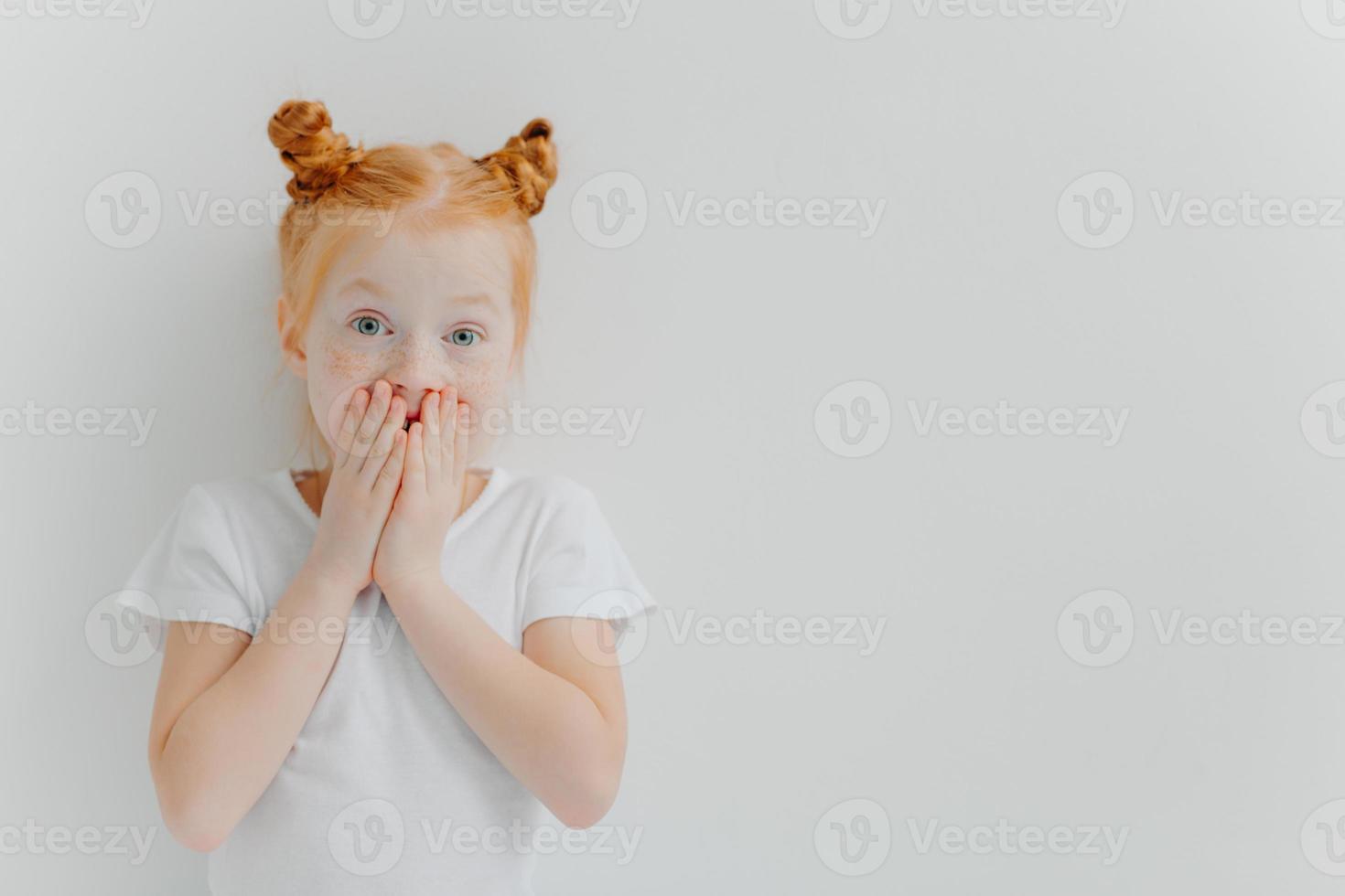 Horizontal shot of surprised red head girl covers mouth, watches exciting cartoon, has unexpected reaction, wears casual white t shirt, stands indoor, expresses wonder. Children and emotions concept photo