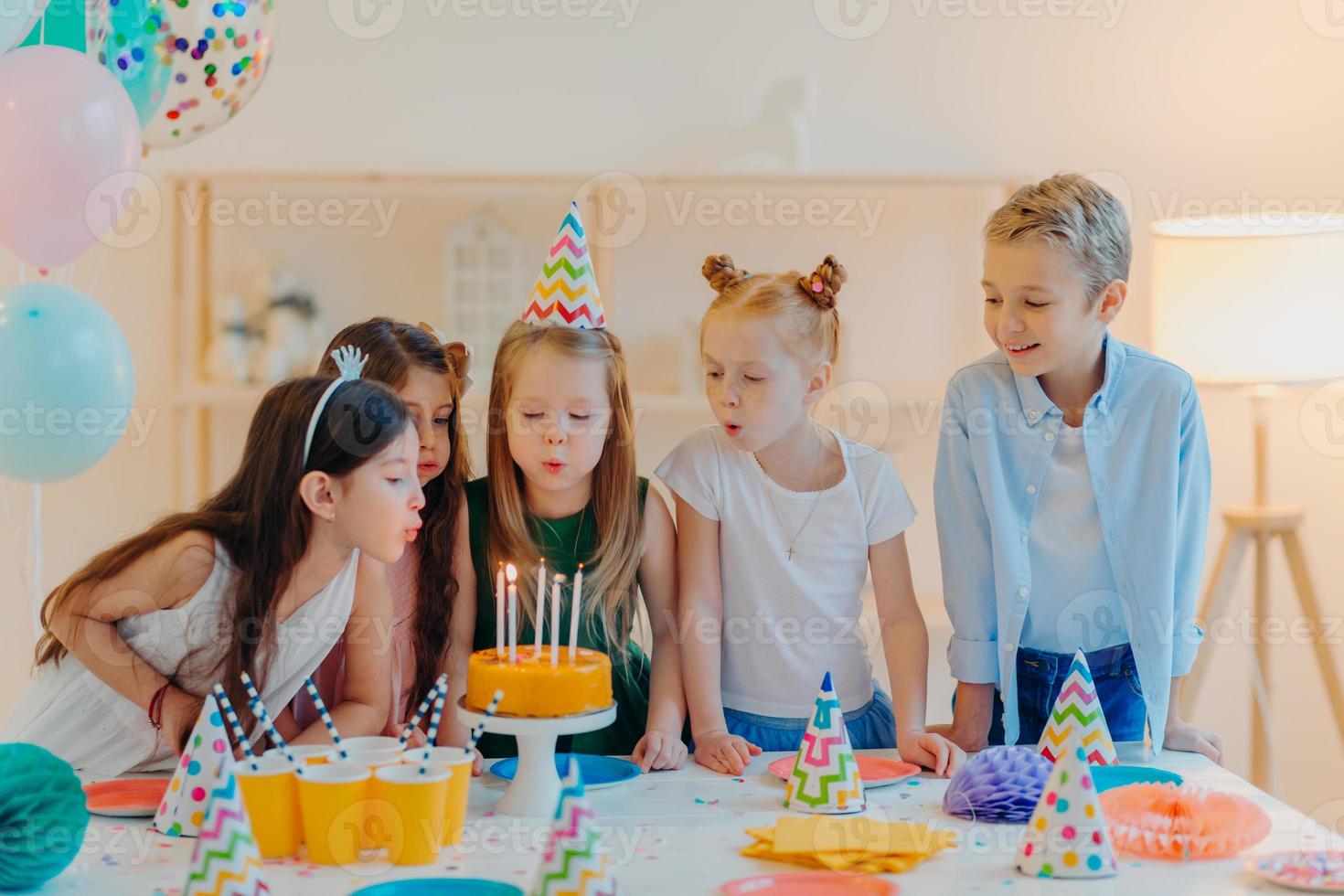 Small kids celebrate birthday party, blow candles on cake, gather at festive table, have good mood, enoy spending time together, make wish, wear party hats, pose indoor with inflated balloons photo