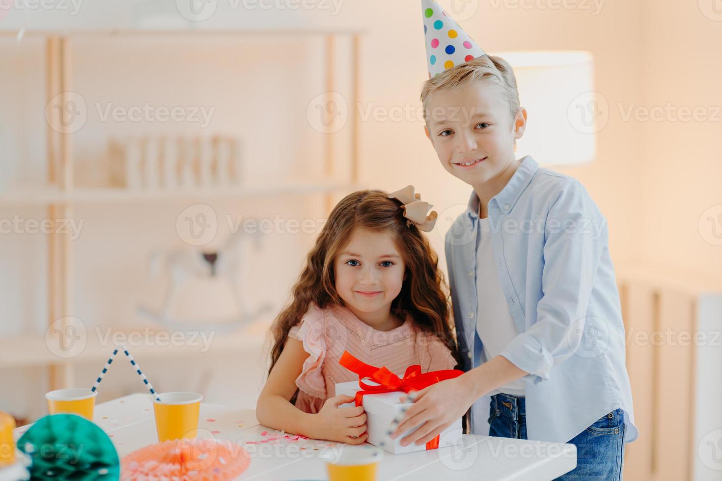 Happy small girl and boy hold gift box with red ribbon, prepare surprise on birthday, pose at white table, wear cone party hats, have positive expressions. Childhood concept photo