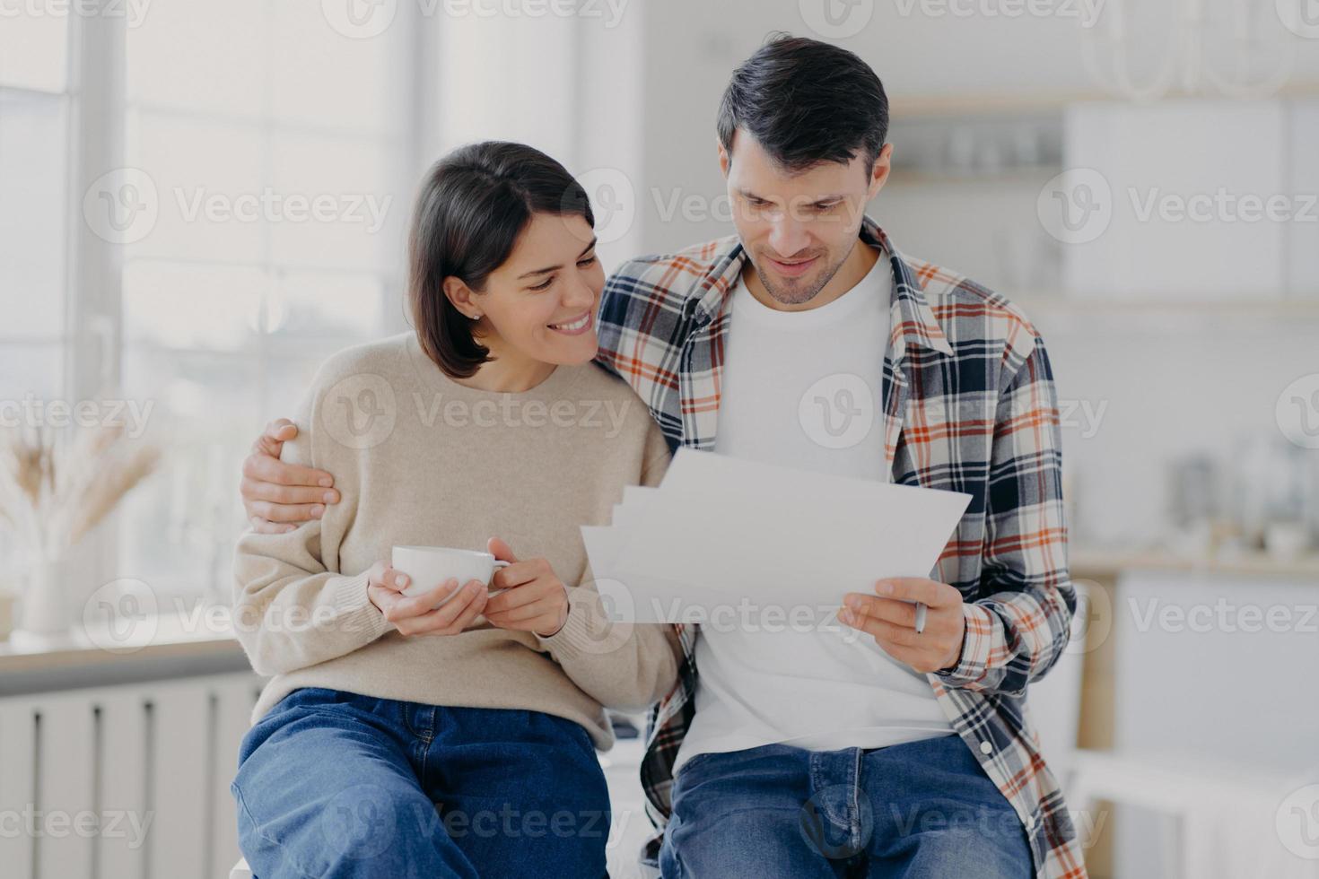 Caring man embraces his wife, look through documents with happy expressions, study payment bills, drinks coffee, dressed in casual wear, pose in spacious room with blurred background. Family and work photo